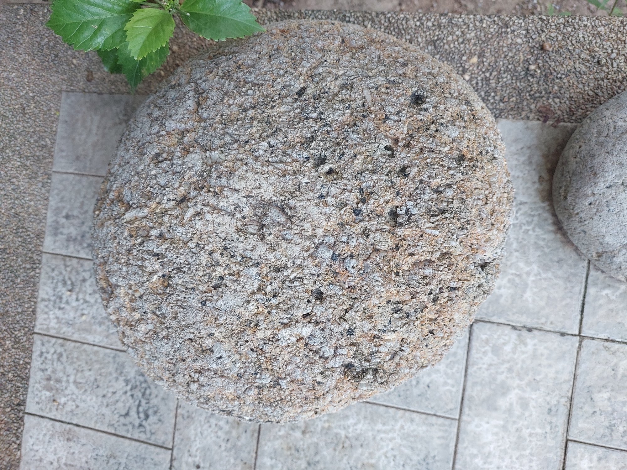 A close-up, overhead shot features a rough, circular stone object resting on a tiled surface. The stone's texture is granular and appears to be made of various shades of gray and tan pebbles embedded in a lighter matrix. The tiling beneath is light gray and arranged in a grid pattern. To the right of the stone, out of focus, are green leaves from a plant. The environment suggests an outdoor setting, possibly a garden or patio area. No people or actions are visible. The location is Baan Mae Nam, Thailand.