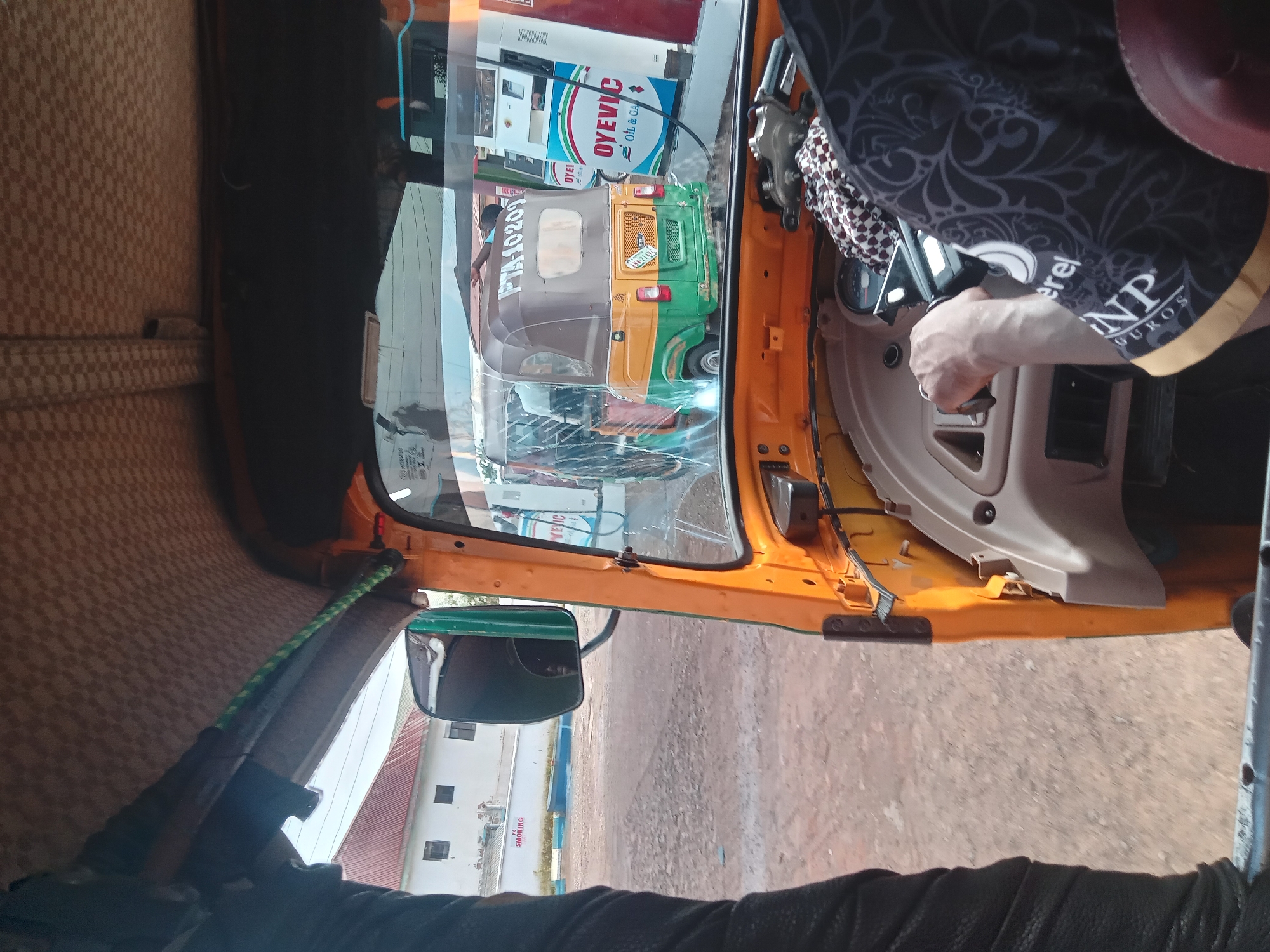 The image is taken from inside an orange auto-rickshaw in Jos, Nigeria, looking forward. The auto-rickshaw has a checkered brown fabric ceiling and a beige dashboard. A person's hands are on the steering wheel. To the right, there is a fuel pump with "OYEVIC Oil & Gas" branding. Another auto-rickshaw, green and yellow, with the license plate "PTA-10209", is parked at the fuel pump. In the background, a white building with a red roof is visible. The ground appears to be unpaved.