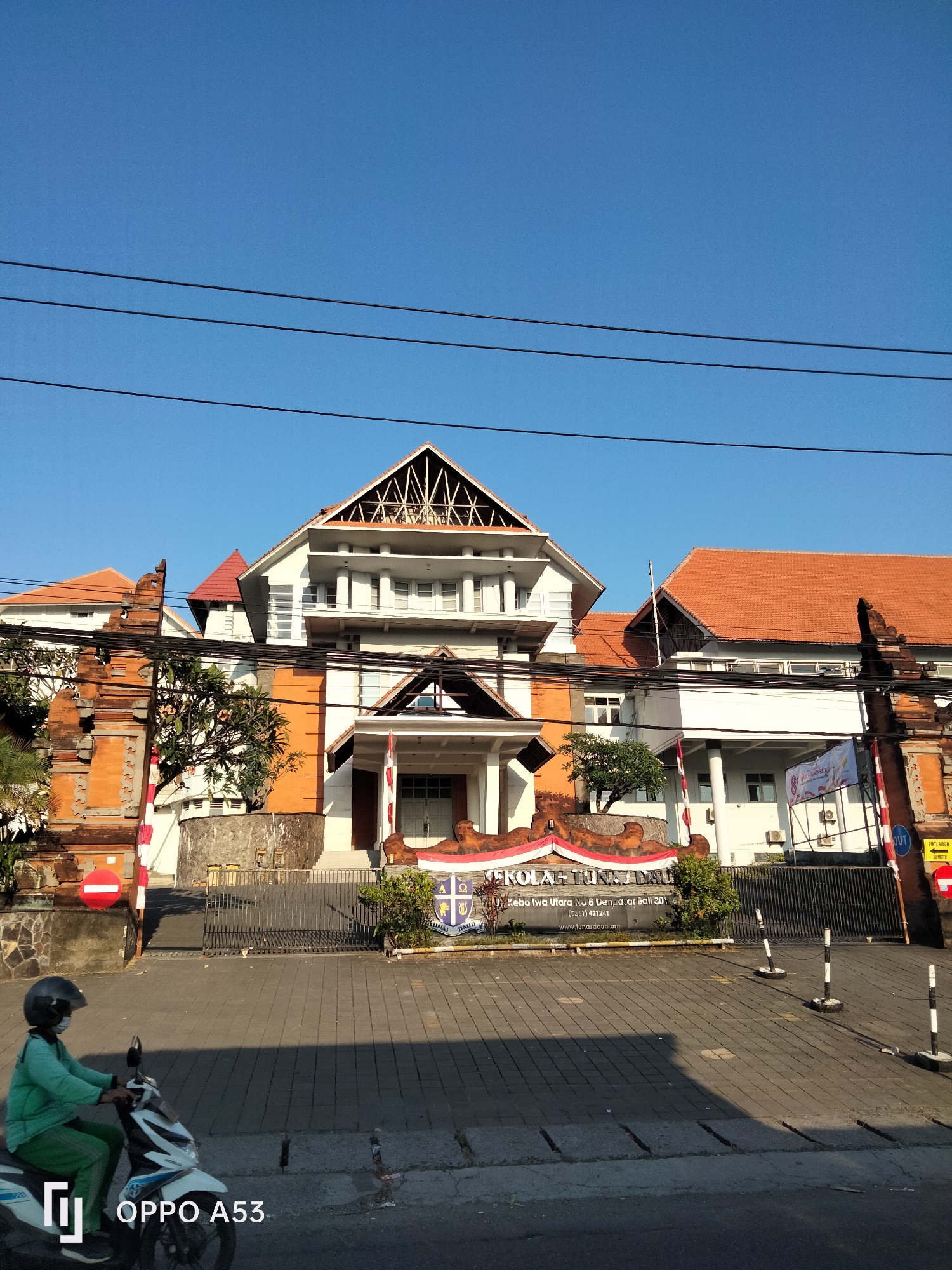 The image shows the front entrance of a school named "SEKOLAH TUNAS DAUD" in Denpasar, Indonesia. The school building features traditional Balinese architectural elements with orange tiled roofs and ornate stone carvings on the gate posts. In the foreground, a person wearing a green uniform and a helmet is riding a white scooter. The street in front of the school is paved with interlocking bricks. The sky is clear and blue. A banner with Indonesian text is displayed at the entrance. The overall scene suggests a sunny day with normal daily activity.