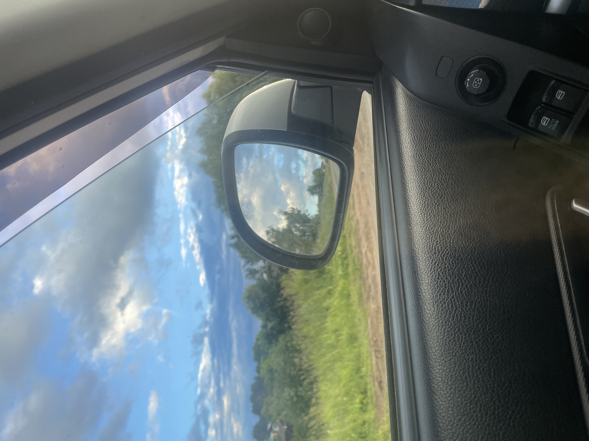 The image is a close-up view from inside a car, looking out the side window and into the side mirror. The scene outside is rural, with green fields and trees under a cloudy blue sky. The side mirror reflects the sky and a line of trees.
Inside the car, the focus is on the door panel. We see the textured black plastic of the door, the window frame, and the edge of the car's interior trim. A side mirror, mounted on the exterior of the car, is visible through the window, and its reflection is captured in the mirror itself. On the door panel, there are control buttons, including a circular dial with directional arrows and two rectangular buttons, one labeled "AUTO DOWN."
The lighting suggests it is daytime, possibly late afternoon, given the soft light and the presence of clouds. The overall impression is one of a quiet moment inside a vehicle, observing the surrounding countryside. There is no discernible text in the image, other than the labels on the window control buttons. The setting appears to be rural Russia, based on the provided location context.