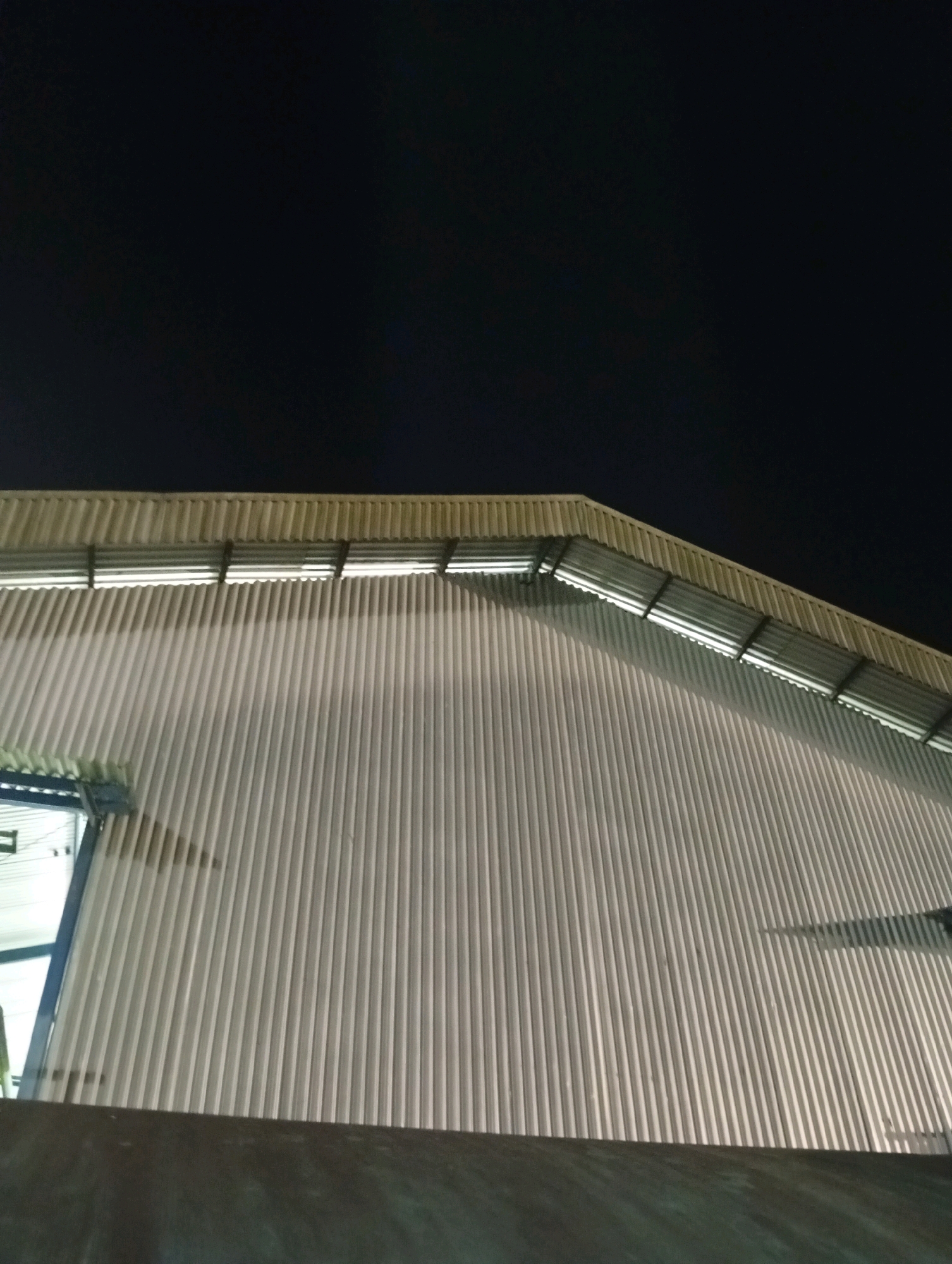 The image shows the exterior of a corrugated metal building at night. The building has a light-colored, ribbed facade. A sloped roof with a metal gutter system is visible at the top of the building. Lights are illuminating the underside of the roof. A dark, possibly shadowed, area is present at the bottom of the image, suggesting foreground elements. The sky above is dark and clear. The scene is generic and does not contain any recognizable landmarks or specific identifying features that would allow for pinpointing a location within Unknown, Indonesia.