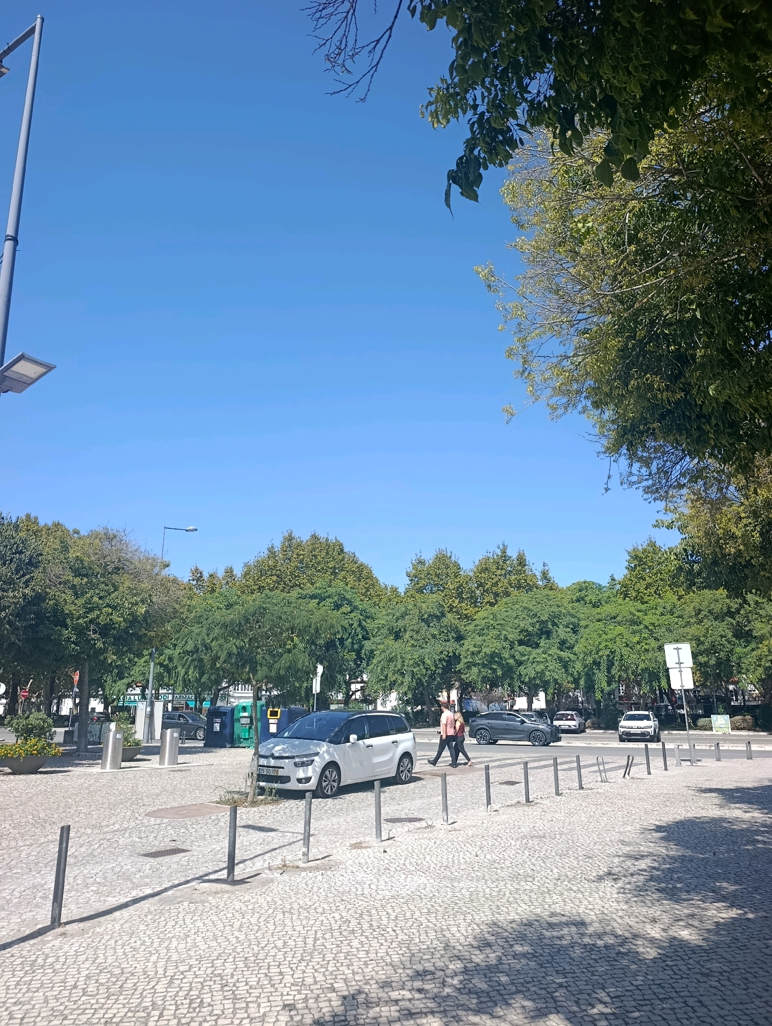 In Setúbal, Portugal, a paved plaza with cobblestone is visible under a clear blue sky. A modern street lamp with a rectangular fixture stands on the left, and bare tree branches are visible in the upper right corner. A line of green trees forms a backdrop across the scene, with some darker foliage on the right side of the image.
In the plaza, a white minivan with a black roof is parked. To its right, two people are walking across the street. A dark gray SUV is driving past them. Further down the street, a white SUV is parked. Several bollards are strategically placed throughout the plaza, some with bicycle racks attached. A sign with a white horizontal bar on top and a white vertical bar below is visible on the right side of the street. The ground is dappled with sunlight and shadow. In the background, buildings and more trees can be seen.