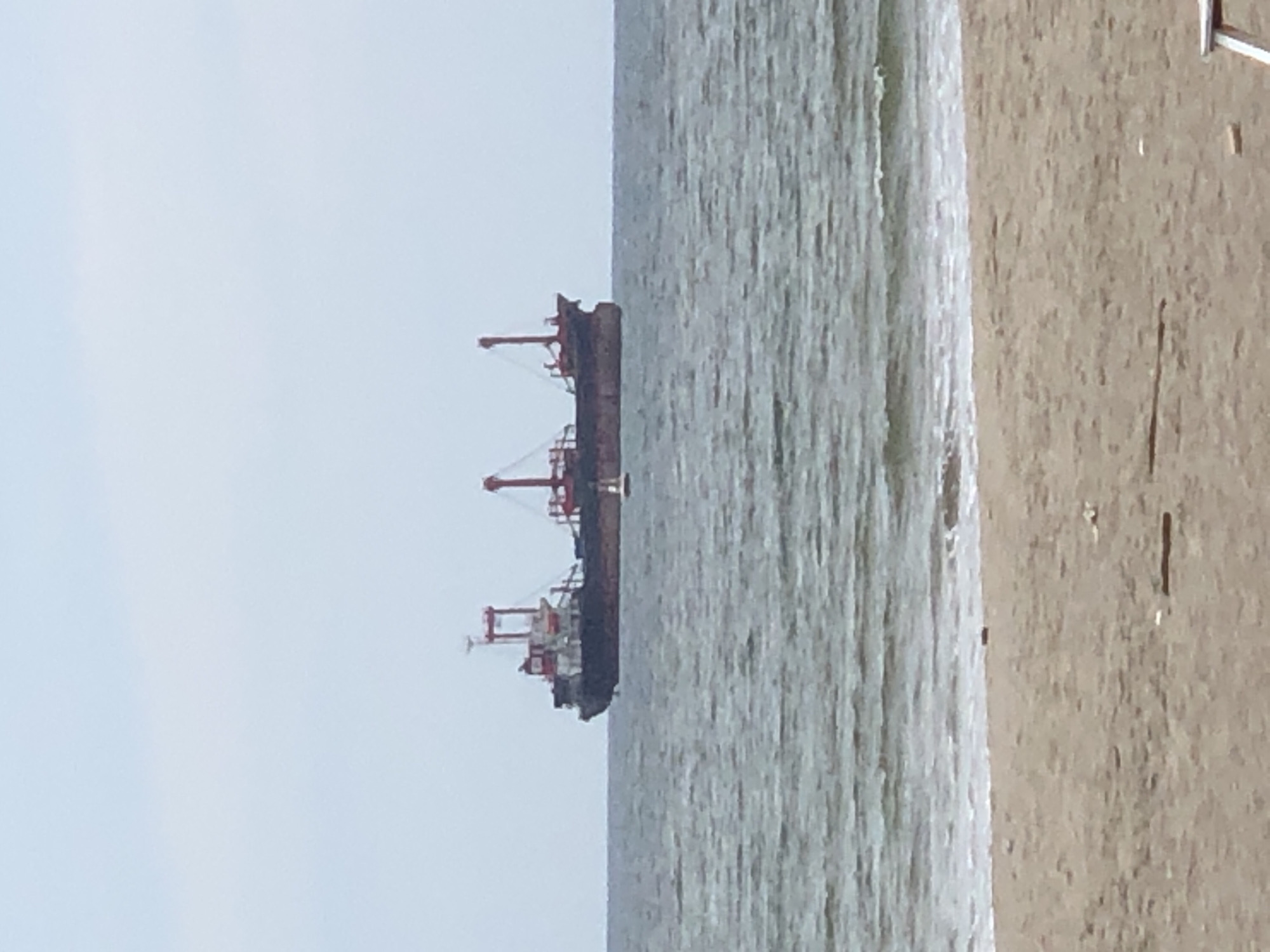 This is a vertical, medium-angle, full shot photograph taken outdoors during the day. The scene depicts a large, dark-colored ship with red cranes docked in the ocean. The ship is positioned in the center of the frame, oriented vertically with its bow to the top. The water is a light gray and appears choppy, with ripples and small waves. To the right of the water is a sandy beach, rendered in a sandy brown color, with some small debris scattered across its surface. The sky, visible on the left side of the frame, is a pale, overcast blue. The overall mood of the image is subdued, likely due to the overcast weather. The image appears to have been taken at a distance, as fine details of the ship and beach are not clearly discernible. The orientation of the photograph is unusual, with the majority of the scene presented sideways. There is no visible text in the image.