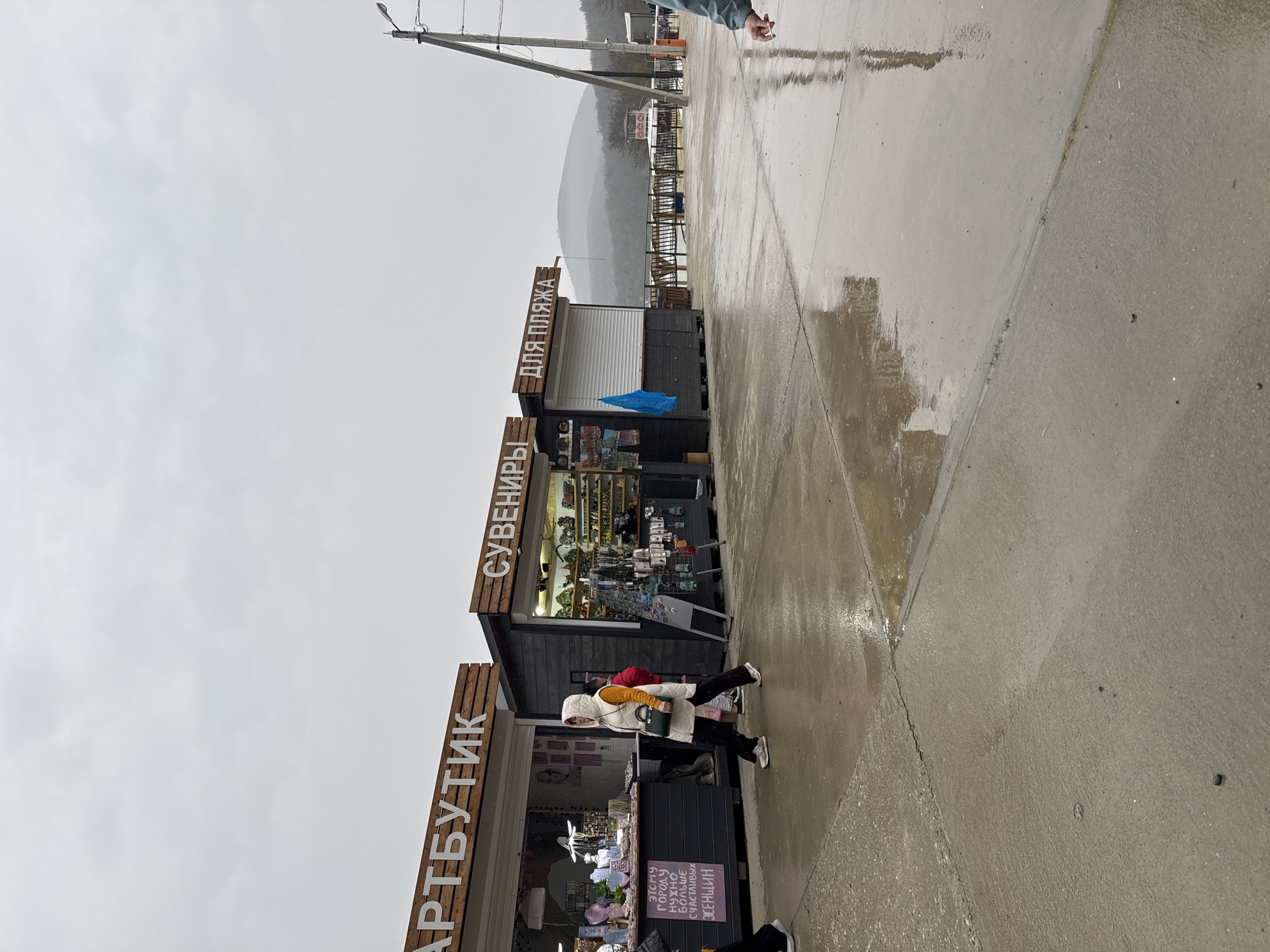 The image depicts a wet, overcast day in Sukko, Russia, along a paved walkway. Three vendor stalls line the walkway, each with signage in Cyrillic: "АРТБУТИК" (Art Boutique), "СУВЕНИРЫ" (Souvenirs), and "ДЛЯ ПЛЯЖА" (For the Beach). A woman wearing a white jacket and carrying a dark handbag walks past the "АРТБУТИК" stall. The walkway is wet, reflecting the surroundings. In the background, power lines and a mountain range are visible through the hazy sky.