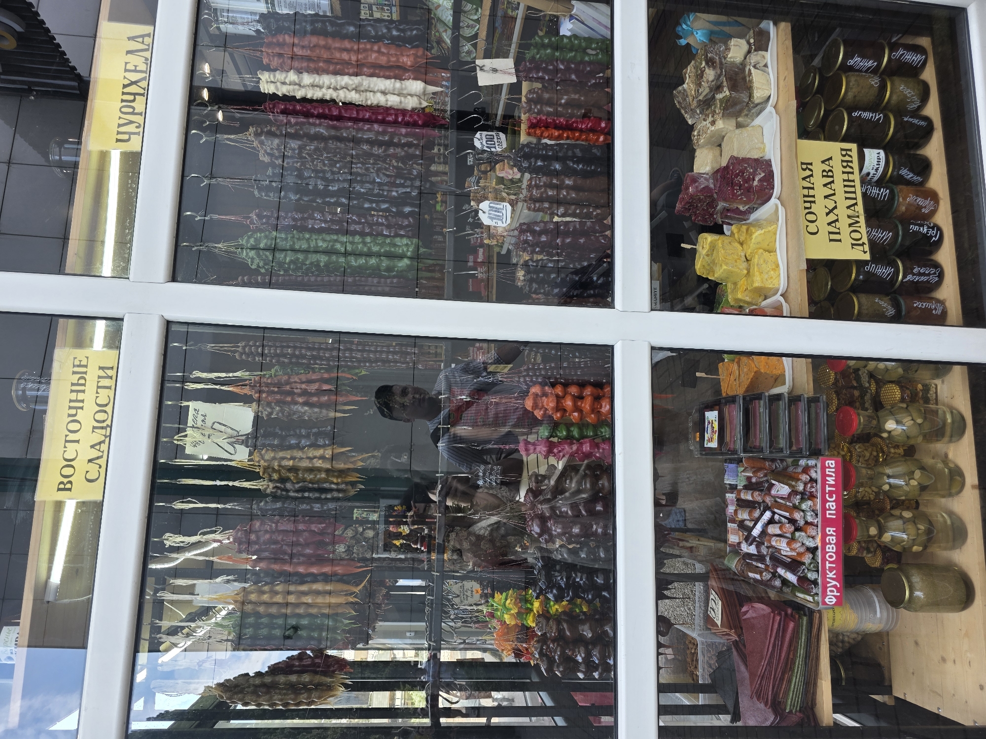 This image captures a vibrant storefront display of various traditional sweets and preserves, likely in Sochi, Russia, given the context and Cyrillic text. The scene is viewed through a large glass window, creating reflections of the outside environment, including a person (possibly a vendor) in a striped shirt and cap, and a clear blue sky with clouds.
The display is divided into several sections. The prominent items are numerous strings of Churchkhela, a traditional Georgian candle-shaped candy made of nuts threaded on a string and coated in thickened fruit juice. These are arranged vertically and horizontally in various colors, including white, red, green, black, brown, and orange. Small price tags showing "100" are visible among them. A yellow sign on the upper left reads "ЧУРЧХЕЛА" (Churchkhela). Below it, another yellow sign states "ВОСТОЧНЫЕ СЛАДОСТИ" (Eastern Sweets).
To the right, another section showcases "СОЧНАЯ ПАХЛАВА ДОМАШНЯЯ" (Juicy Homemade Pahlava), with pieces of varying shapes and colors, including yellow, red-brown, and white-beige varieties, neatly arranged in trays. Adjacent to the pahlava are rows of glass jars filled with preserves or jam. Visible labels on the jars include "ИНЖИР" (Fig), "Фейхоа" (Feijoa), "Абрикос" (Apricot), "Грецкий" (Walnut), "Белая" (White), "Варенье" (Jam), "шиповник" (rosehip), and "АЖИРА".
The lower right section features "Фруктовая пастила" (Fruit Pastila), displayed both as rolled tubes and flat sheets in different colors, next to more jars of preserves. Another product resembling nuts or small fruits in syrup is also seen in jars. The overall impression is a well-stocked shop, presenting a rich variety of local or regional delicacies under clear, daytime conditions.