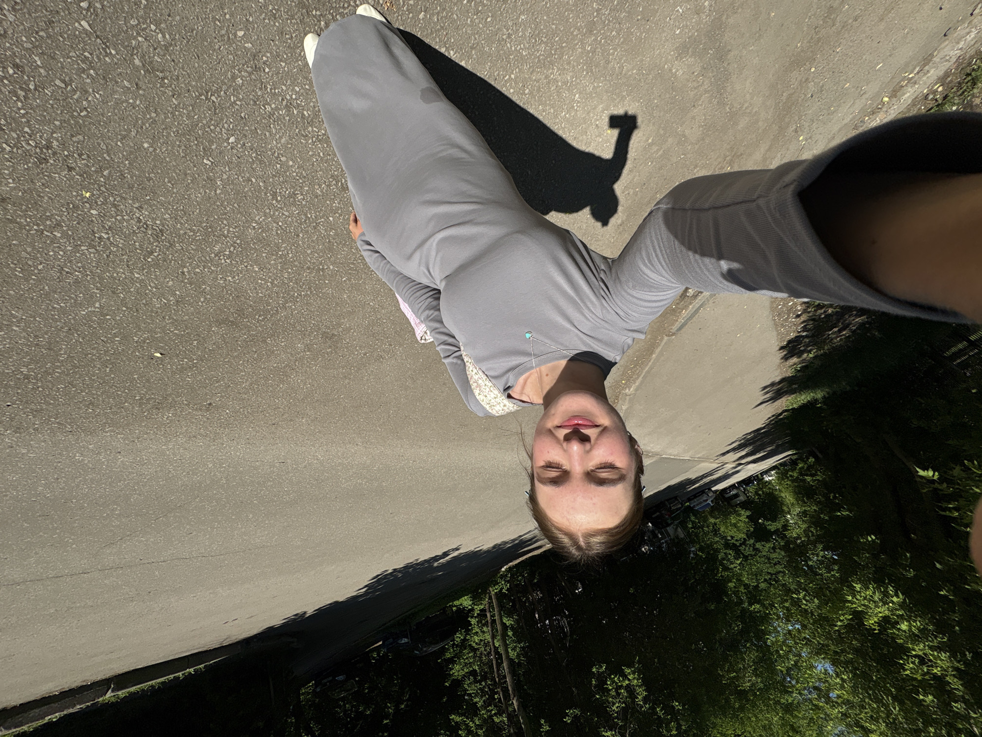 A woman stands on an asphalt path in Perm, Russia, likely taking a selfie. She is wearing a long, gray dress and white shoes. Her shadow is cast on the path next to her. To her right, a line of trees is visible, and a road or parking area with vehicles. The lighting indicates it is likely daytime.