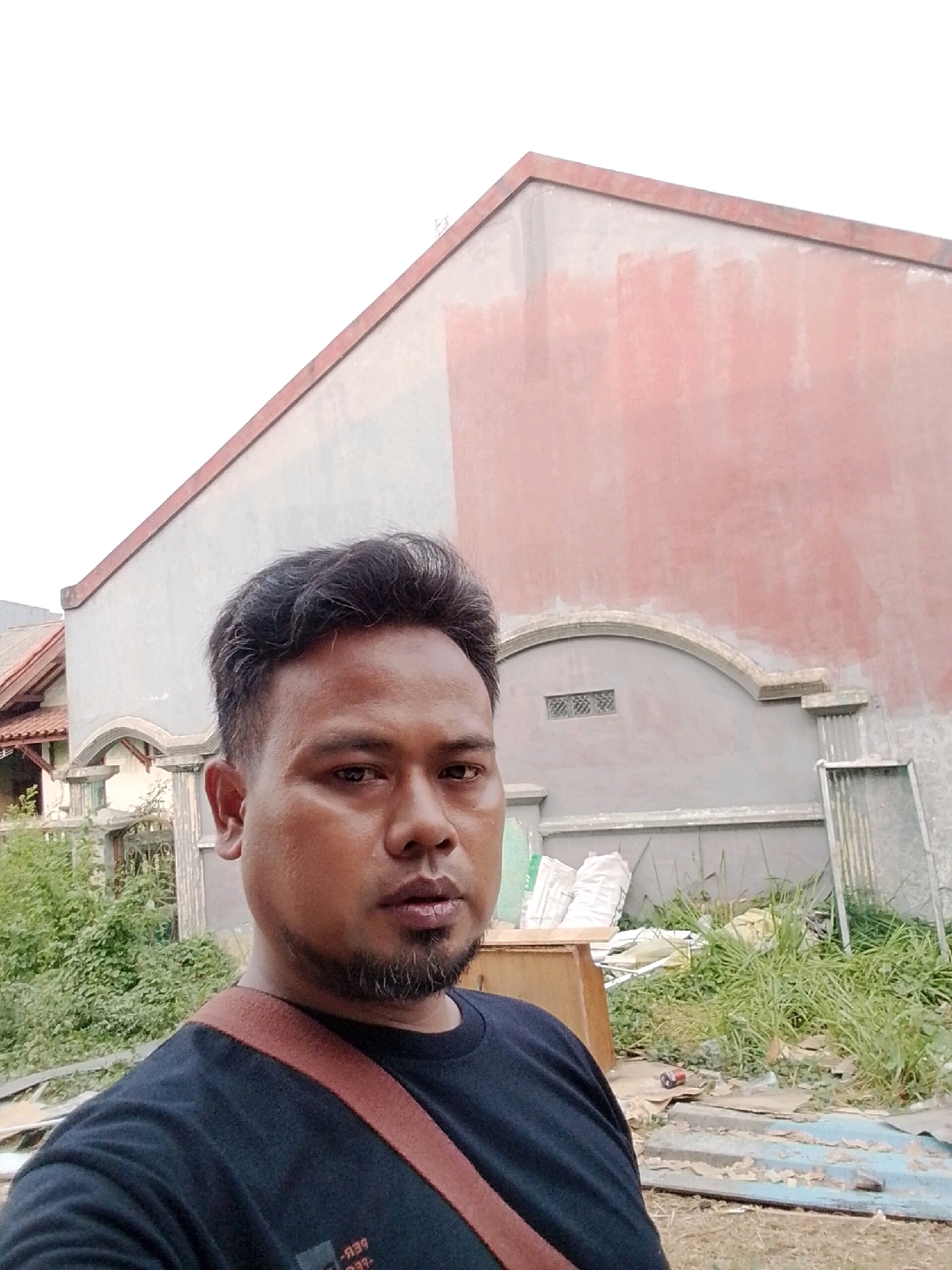 In the city of Babelan, Indonesia, a man with dark hair and a goatee is taking a selfie outdoors. He is wearing a black t-shirt with a reddish-brown strap across his chest. Behind him, a building with a red and gray facade is visible. The area appears to be under construction or renovation, with building materials and overgrown vegetation present. The sky is overcast.