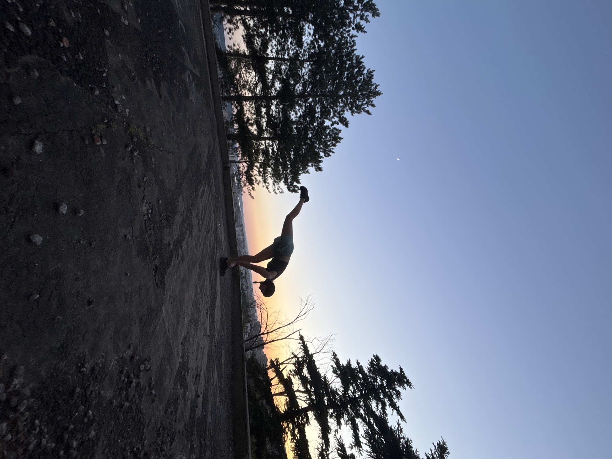 A silhouette of a person is performing a yoga pose on a dark, rough surface, possibly a rooftop or abandoned area, with trees and a city skyline in the background. The sky is a clear gradient from a deep blue at the top to a soft orange and pink near the horizon, indicating sunset or sunrise. A crescent moon is visible in the upper left portion of the sky. The person is positioned in the center of the frame, bending forward with one leg extended backward and upward, creating a dynamic and balanced posture. The trees, silhouetted against the bright sky, are evergreen and appear to be tall and slender. The ground is textured with small stones and debris, suggesting an unmaintained or natural outdoor space. The overall scene evokes a sense of tranquility and physical exertion against a natural backdrop.