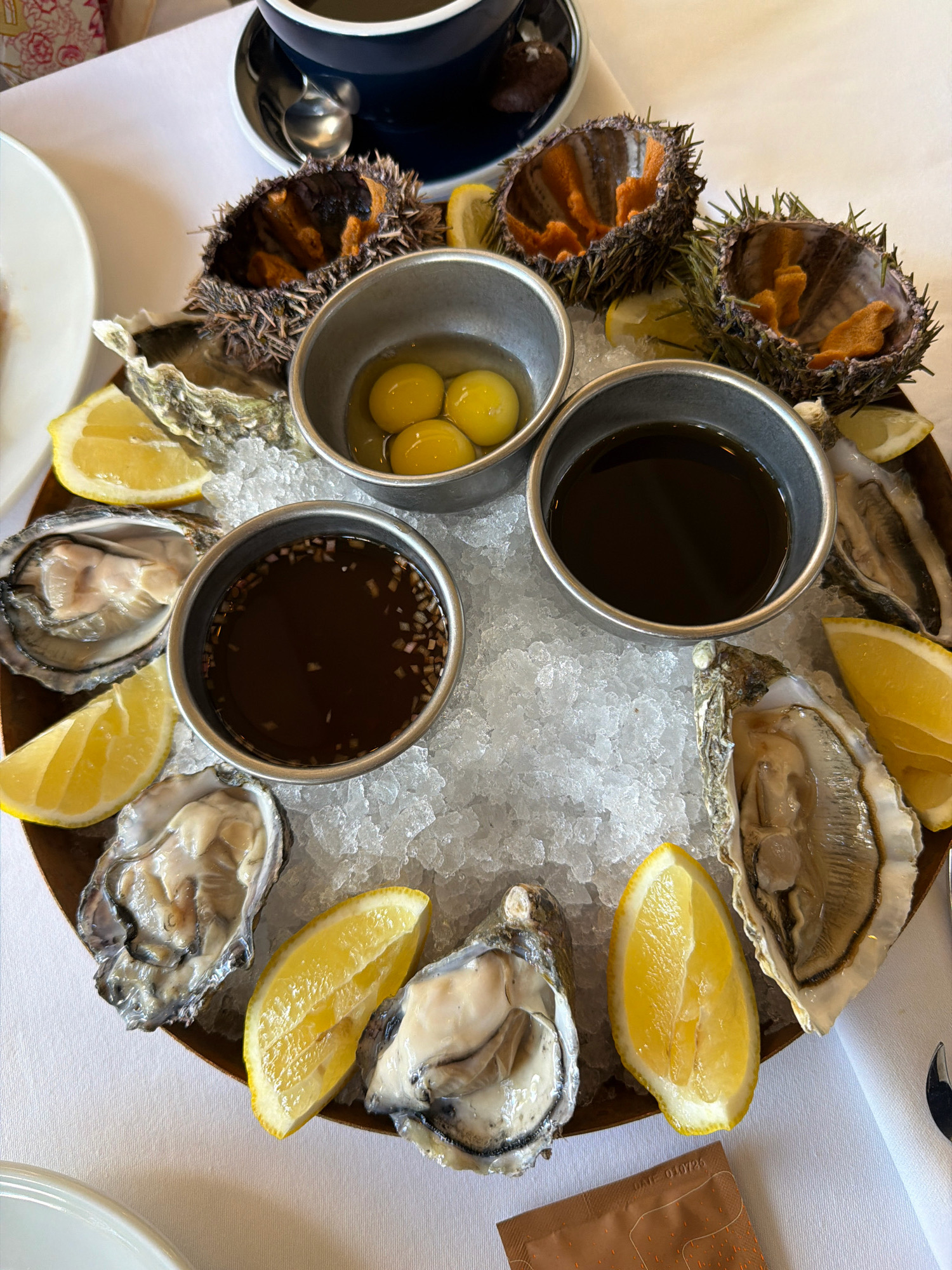 The image displays a seafood platter served on a wooden tray filled with crushed ice. The platter features several oysters, lemon wedges, and two small metal bowls. One bowl contains three egg yolks, while the other holds a dark liquid, possibly a sauce or mignonette. In the background, there are sea urchins in their shells, also arranged on ice. A dark blue cup and saucer with a spoon are visible on a white tablecloth. The scene appears to be set in a restaurant, and there is no indication of the city or country where the photo was taken.