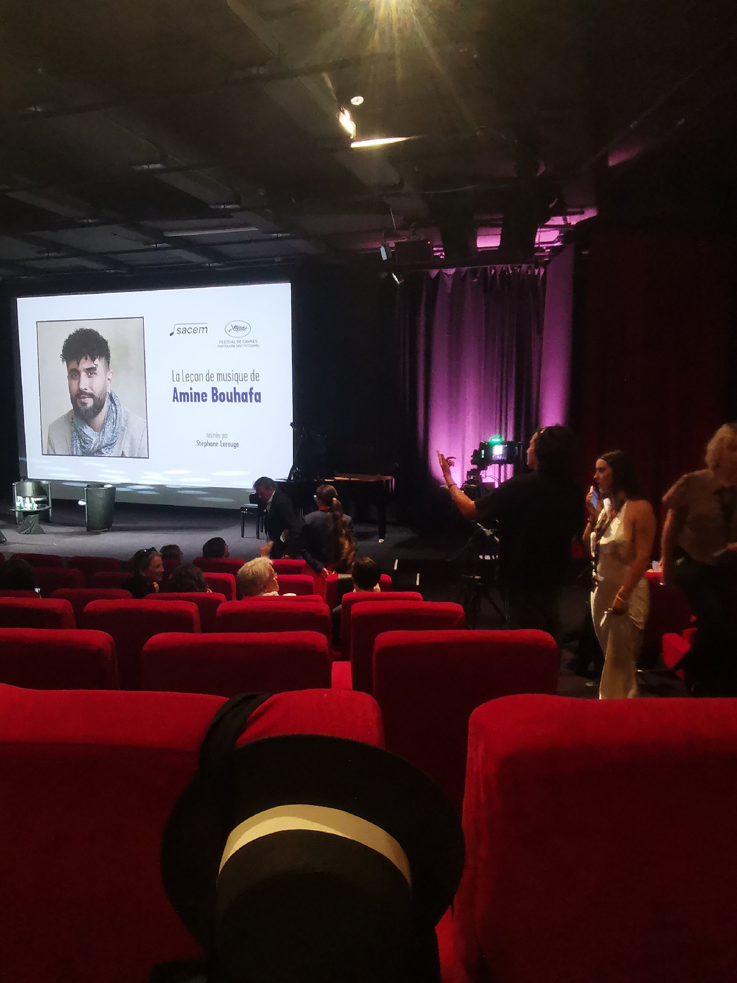 The image captures a scene inside a theater, possibly in Cannes, France, as suggested by the "Festival de Cannes" logo on the screen. The focus is on a presentation titled "La Leçon de Musique de Amine Bouhafa," which translates to "The Music Lesson of Amine Bouhafa." A portrait of Amine Bouhafa is displayed on the screen alongside the logo of Sacem, a society of authors, composers, and music publishers. The presenter is noted as Stéphane Lerouge.
The theater has red cushioned seats, typical of cinemas or auditoriums. In the foreground, a black top hat with a white band is visible, partially obstructing the view of the seats. Several people are seated or standing in the middle ground, including an individual adjusting equipment, possibly for recording or filming the presentation. A piano is positioned on the stage behind the seated individuals. The lighting is dim with pink accent lights illuminating the curtains in the background. The general setting suggests a special event or screening related to music and film, possibly during the Cannes Film Festival.