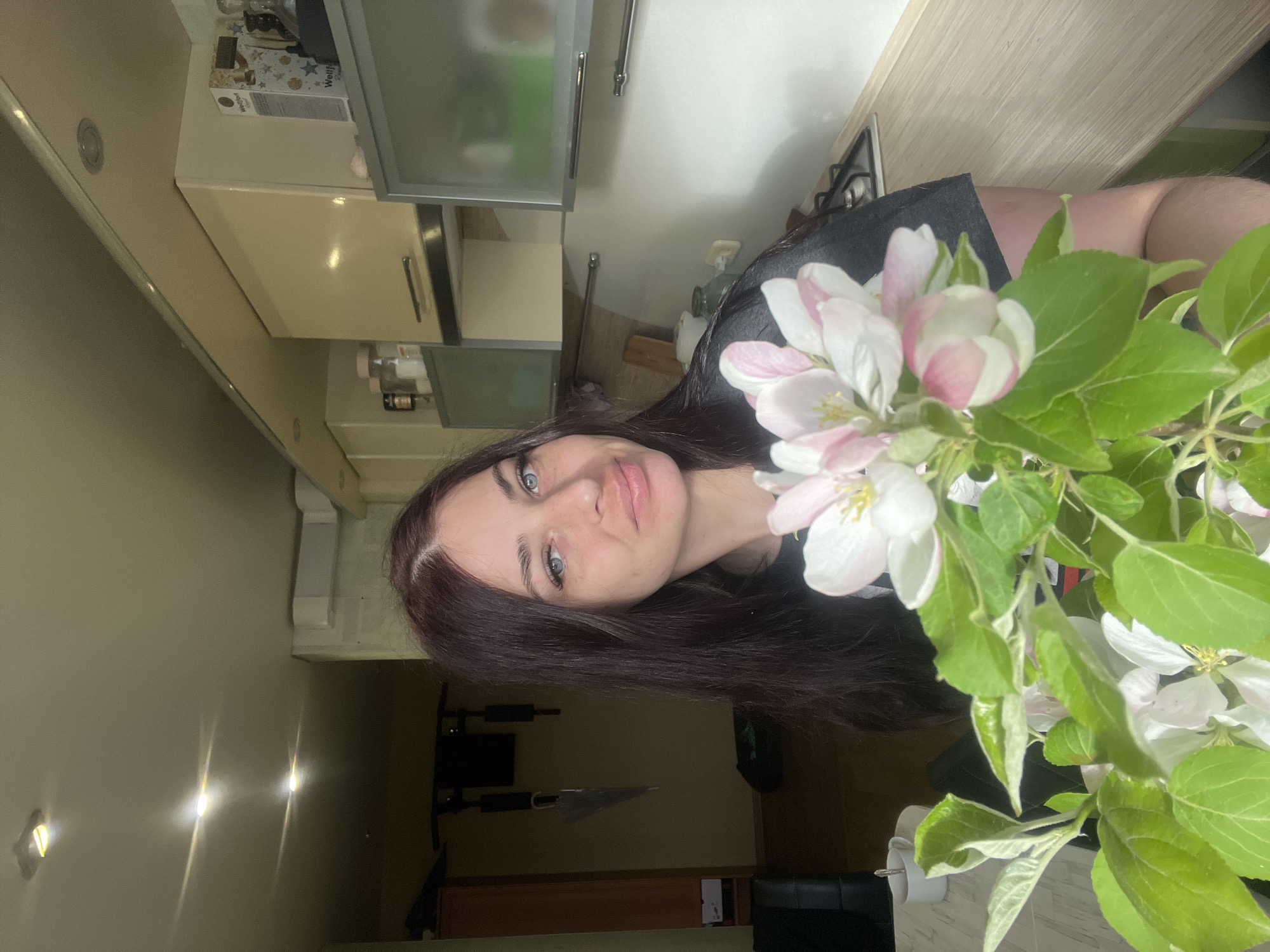 A woman with fair skin, blue eyes, and dark brown hair is centered in the image, holding a vase or bunch of flowering branches. The flowers are white and light pink. The background suggests an interior setting, likely a kitchen, with light-colored cabinets and a countertop. A glimpse of a living area with a TV and furniture is visible in the lower part of the image. The lighting appears to be a combination of natural and artificial light sources. The location is possibly Chaikovsky, Russia, but this is only implied, not visually verifiable.