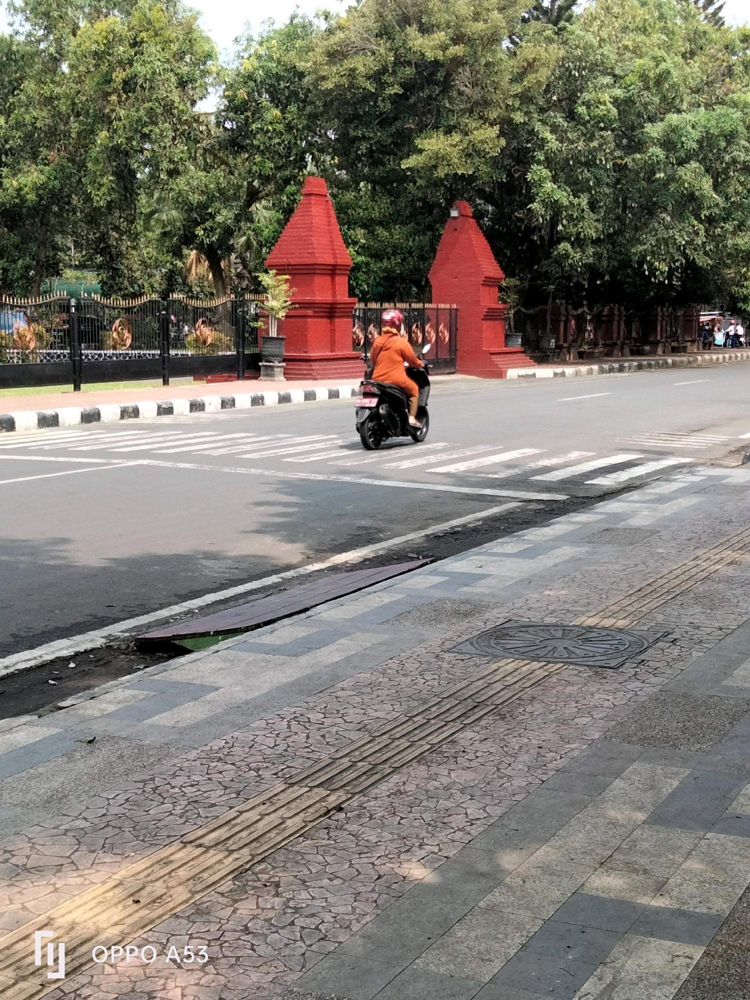 In Trenggalek, Indonesia, a person wearing an orange jacket and a red helmet rides a motorcycle across a crosswalk on a city street. The road is paved and marked with white stripes for the pedestrian crossing. To the left of the road, a black metal fence with gold accents encloses an area with lush green trees and two distinctive red, pyramidal structures. A drain grate is visible on the sidewalk on the right side of the road. The sky is overcast, casting shadows on the pavement. The overall scene depicts a typical urban environment in Indonesia.