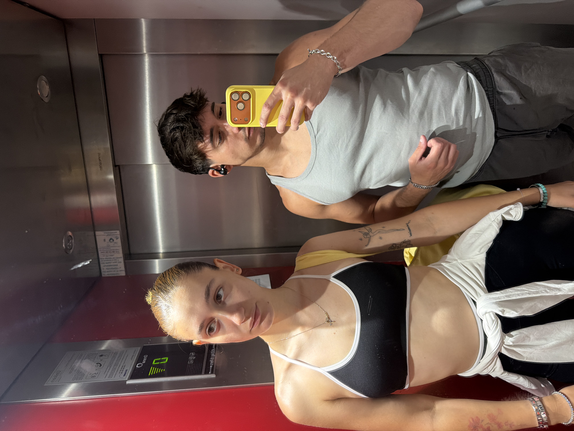 An eye-level photograph depicts two young adults inside an elevator, capturing a mirror selfie. The elevator's interior features a red wall on the left, which has a reflective panel displaying reversed text including "Първомайски" (Pŭrvomayski), identifying the location as Sofia, Bulgaria. Below this text, a digital display shows the number "0" with a green upward arrow. The remaining walls of the elevator are constructed from brushed metal panels.
On the left, a female subject with light brown hair pulled back wears a black sports bra with white trim, black leggings, and a white long-sleeved top tied around her waist. She has a gold-colored necklace with a small charm, multiple bracelets on her left wrist, and a turquoise bracelet on her right wrist. A reddish floral tattoo is visible on her left forearm, and a stylized outline tattoo of a figure is present on her right forearm. She is looking slightly to her right, away from the camera's direct line of sight.
On the right, a male subject with short, dark, wavy hair and faint facial hair wears a light gray ribbed tank top and dark gray athletic shorts with a drawstring. He has black wireless earbuds in both ears and a silver chain bracelet on his left wrist. He is holding a bright yellow smartphone with a triple-lens camera array in his right hand, positioned to capture the reflection. He is looking down at the phone.