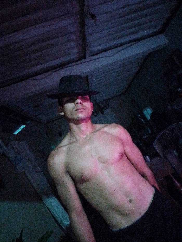 The image depicts a young adult male positioned centrally within a dimly lit, possibly indoor or sheltered environment in Juan Griego, Venezuela. The individual wears a black fedora-style hat, casting a shadow over his eyes, and is shirtless, revealing a muscular torso with defined pectorals and abdominal muscles. He wears dark-colored trousers, of which only the upper edge is visible. The subject is oriented towards the camera, maintaining a direct gaze.
The background reveals a textured ceiling or roof structure, possibly constructed from corrugated material or wooden panels. To the left of the subject, a horizontal light source emitting a cool blue-green illumination is partially visible. To the right, a dark, circular object with a mesh or spoke pattern, consistent with a fan, is partially discernible. The scene is illuminated by multiple light sources: a prominent reddish-pink light highlights the subject's chest and lower face from the front or below, while a cooler, bluish light accents elements of the background and the subject's left side. No specific interactions or significant actions beyond posing for the photograph are evident.