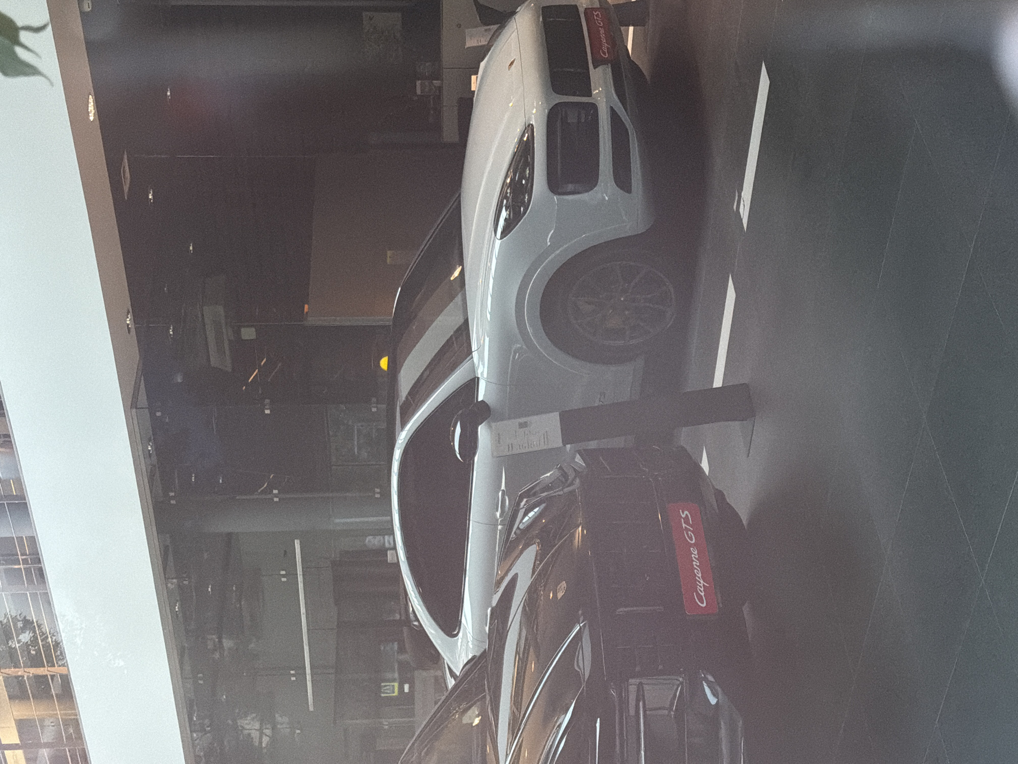 Two Porsche Cayenne GTS vehicles are parked indoors, possibly in a showroom, given the clean floor and modern architecture. The car in the foreground is black, and a red badge on its rear quarter panel clearly reads "Cayenne GTS". The car behind it is white, and a similar red badge is visible on its front fender. Both cars appear to be in pristine condition. The background shows reflective surfaces and building elements, suggesting an indoor, well-lit environment.