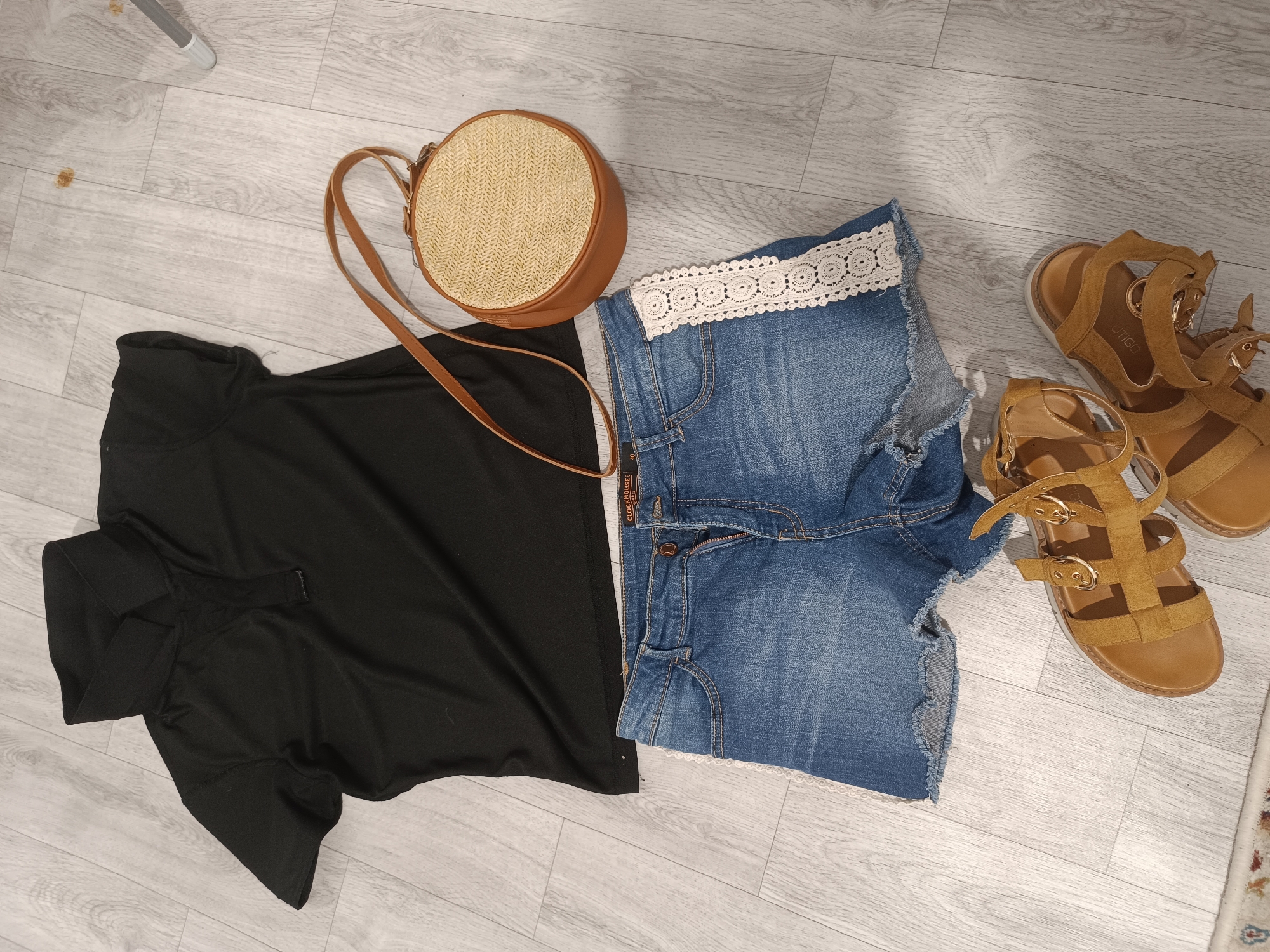 An overhead, eye-level shot captures a flat lay of women's clothing and accessories arranged on a light grey, weathered wood-plank floor. The items are positioned in the lower half of the frame, creating a casual, outfit-planning aesthetic.
On the left, a black, short-sleeved top with a collar lies spread out. To its right, a pair of distressed, medium-wash denim shorts are laid out, featuring a white lace trim at the hem. A small, round straw purse with a brown leather strap is situated above the top and shorts, with the strap draped across them. On the right side of the frame, a pair of tan, strappy gladiator sandals are placed, one behind the other. The flooring extends throughout the image, showing subtle wood grain and color variations. A portion of a patterned rug is visible at the bottom right corner. The scene is well-lit, casting soft shadows and highlighting the textures of the items. There are no people present in the image. The setting is a domestic indoor space. The image does not contain any elements that can be identified with the city of Taraz city administration, Kazakhstan.