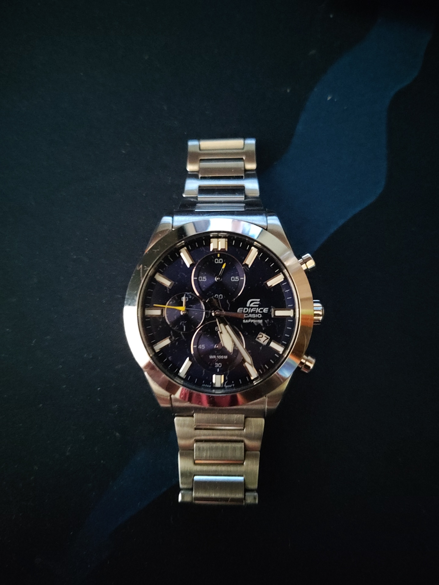 The scene depicts a single Casio Edifice chronograph wristwatch, model "Sapphire," centrally positioned against a dark, matte background in an indoor setting within the city of Plovdiv, Bulgaria. The watch features a stainless steel case with an octagonal bezel and a matching three-link metal bracelet. Its dark blue or black dial includes three sub-dials—a 0.0/0.5 indicator at the 12 o'clock position, a 60-second counter at 9 o'clock, and a 45/30 minute counter at 6 o'clock—along with a date window displaying "27" at the 4 o'clock position. Luminous baton hour markers are present, and the silver-toned hour and minute hands point to approximately 8:56. A distinct yellow hand is visible on the 60-second sub-dial, and the main chronograph second hand has a yellow tip. The dial also bears text indicating "EDIFICE," "CASIO," "SAPPHIRE," "WR 100M," and "JAPAN MOVT." The watch case has two polished pushers and a knurled crown on its right side. Minor superficial scratches are discernible on the bezel near the 10 o'clock mark. The dark background surface exhibits subtle texture and scattered dust specks, with an irregular, lighter blue area visible towards the upper right, possibly a reflection or a distinct surface feature. No people, active actions, or specific interactions are depicted.