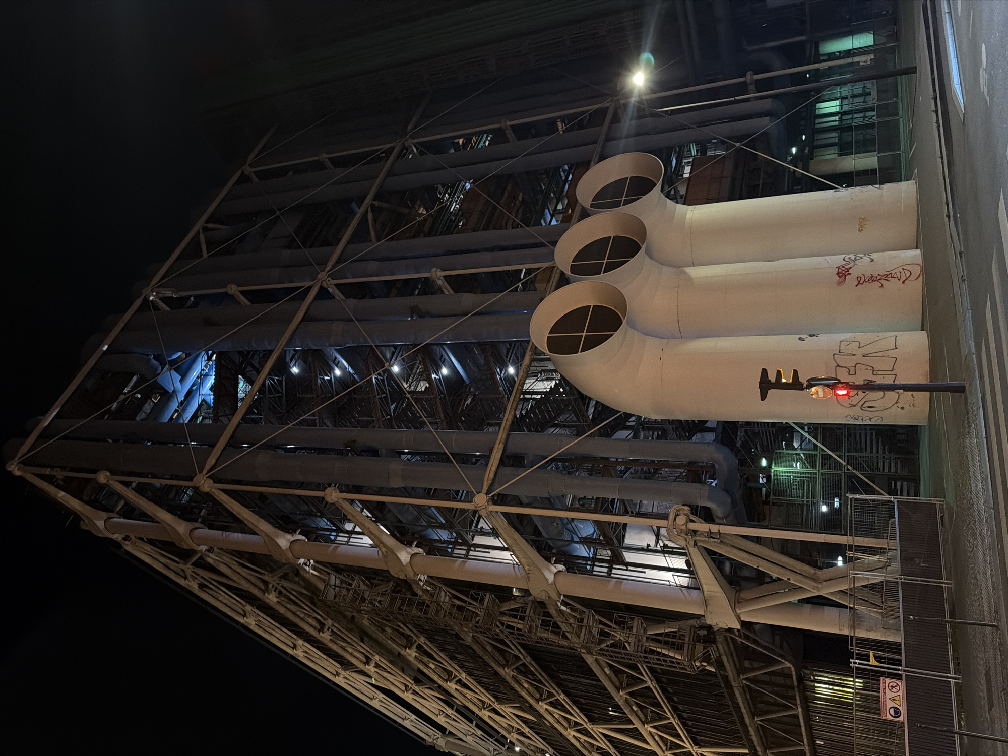 This image depicts the Centre Georges Pompidou, a modern art museum in Paris, France. The photo is taken at night, showcasing the building's distinctive exposed infrastructure, including large pipes and a complex network of metal beams and supports. A traffic light is visible on the right, displaying a red light. Graffiti is visible on the pipes. The image is taken from a low angle, emphasizing the height and industrial aesthetic of the structure.