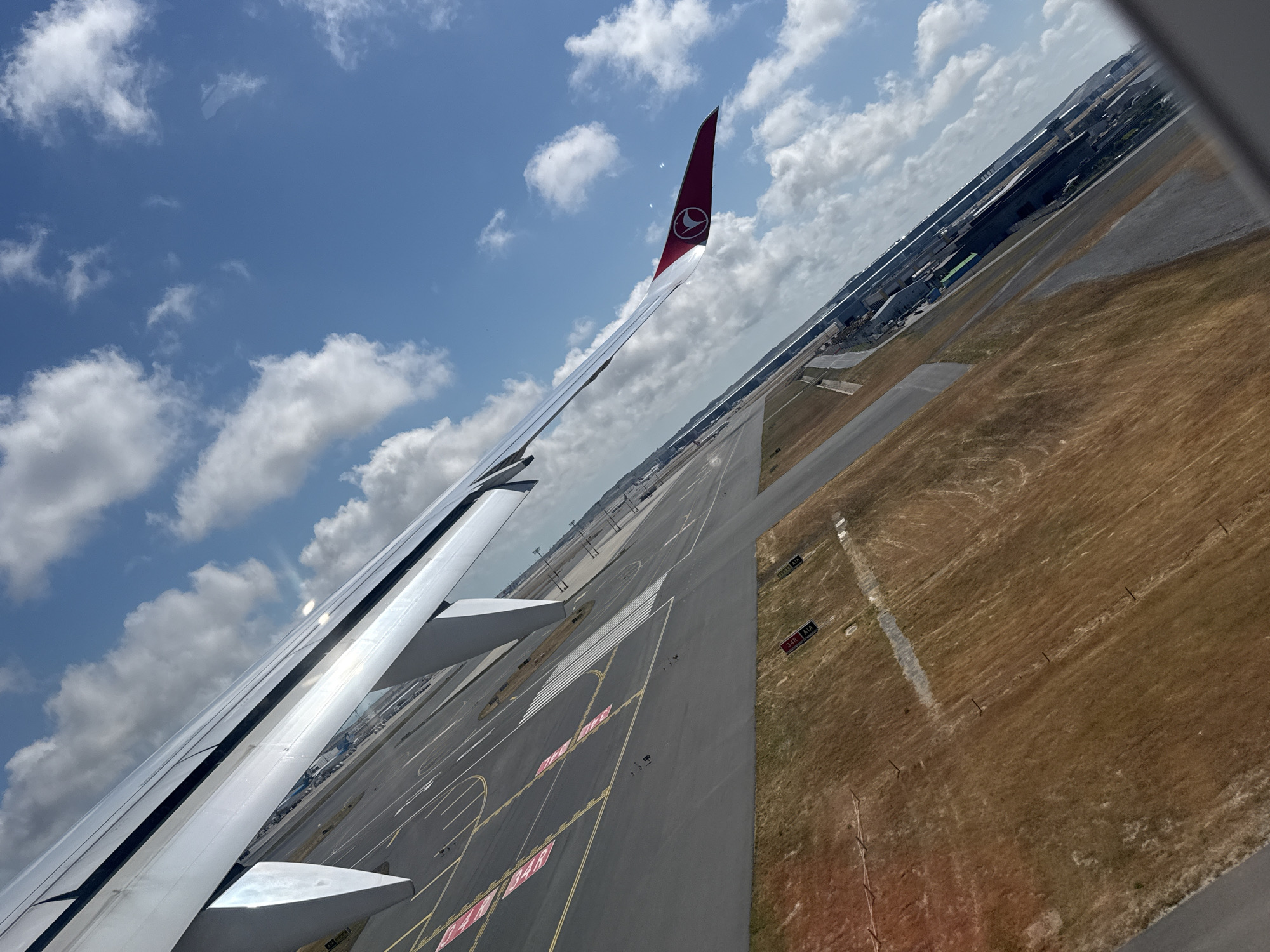 The image, taken from an airplane window, offers a perspective of an airport and its surroundings in Arnavutköy, Turkey. The foreground is dominated by the airplane's wing, gleaming in the sunlight, with its distinctive Turkish Airlines logo visible on the wingtip. Below the wing, the airport tarmac stretches out, marked with various lines and signals, suggesting active runways and taxiways. Beyond the tarmac, dry, brown fields with some scattered vegetation are visible, indicating a semi-arid or seasonal landscape. In the distance, a sprawling urban landscape with numerous buildings and structures can be seen, likely part of the airport infrastructure or the city itself. The sky is a vibrant blue, dotted with fluffy white clouds, suggesting clear weather conditions. The angle of the photograph suggests the plane is either taking off or landing.