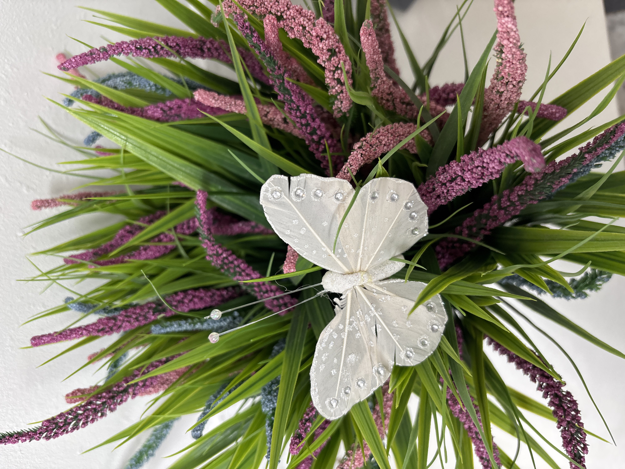 The image presents a close-up view of a floral arrangement. Dominating the scene is a decorative white butterfly with glittering accents positioned centrally. The butterfly is nestled amongst bright green foliage and spikes of mauve and light blue flowers. The background appears to be a plain white surface. The location of Izhevsk, Russia is not discernible from the image content.