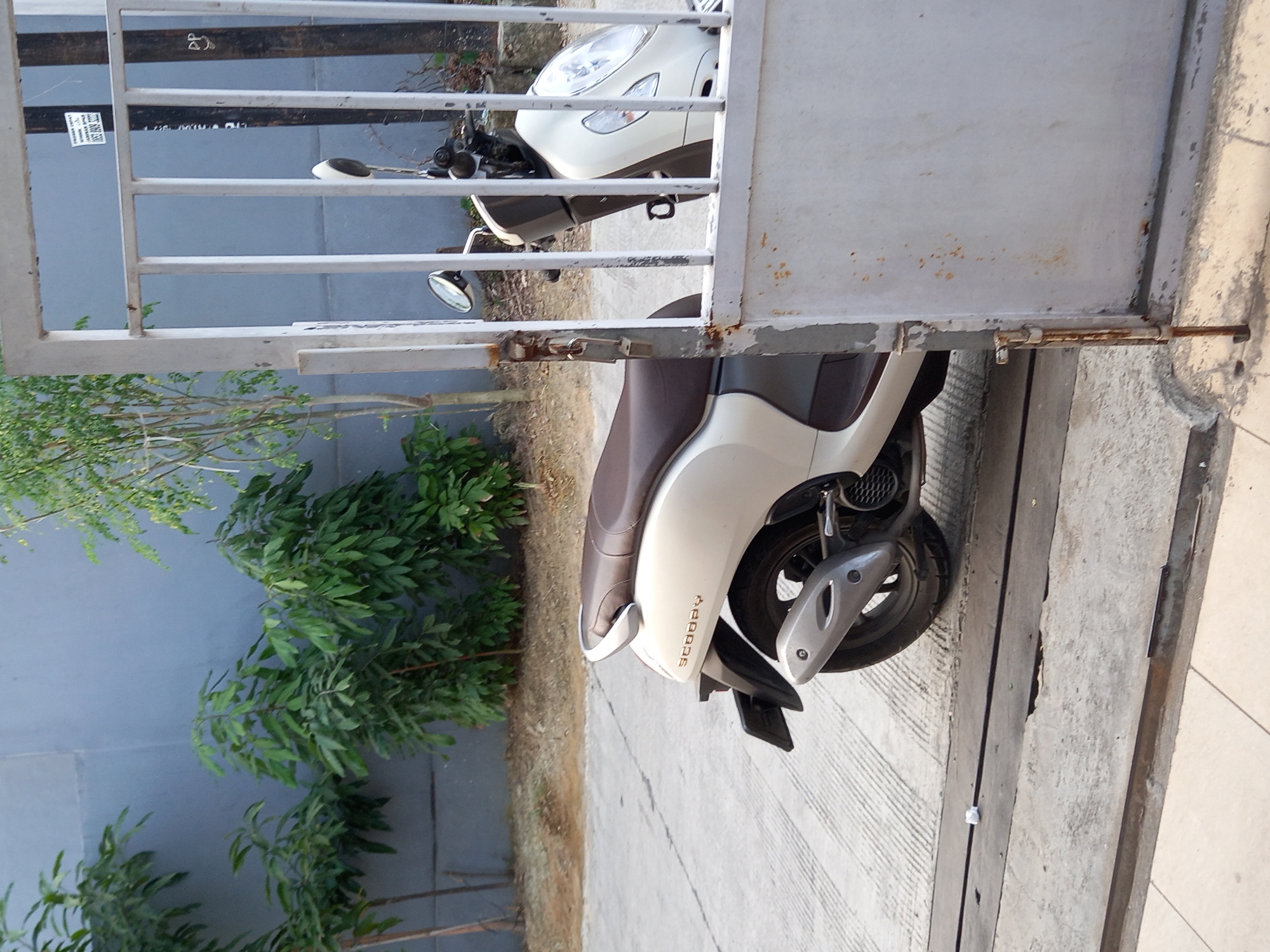An exterior, eye-level shot captures a white scooter parked on a concrete surface, partially obscured by a white metal gate. The scooter is facing left, with its rear and left side visible. It has a brown seat and a silver exhaust pipe. The gate, with vertical bars, is slightly ajar, revealing more of the scooter and some vegetation behind it. The background consists of a grey wall and trees. The scene appears to be located in Samarinda, Indonesia, given the context.