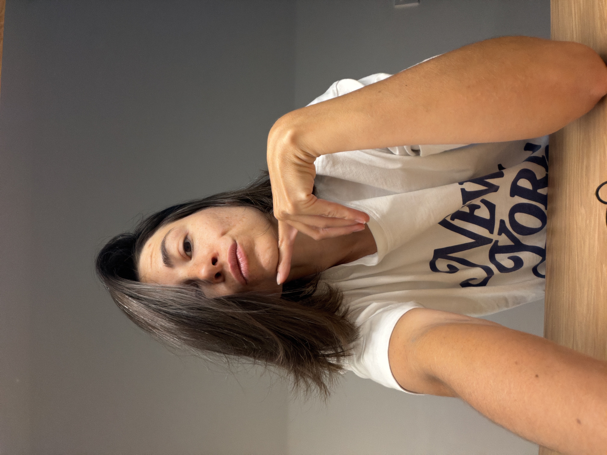 A woman poses for a selfie indoors, possibly in a residence, as the backdrop shows a grey wall on the left. The woman has dark hair and is wearing a white t-shirt with "NEW YORK" printed on it in blue, partially visible on her chest. She has her arm raised and her fingers are near her face. A wooden surface is visible to the right of the woman. There is no explicit indication that the location is Budva, Montenegro.