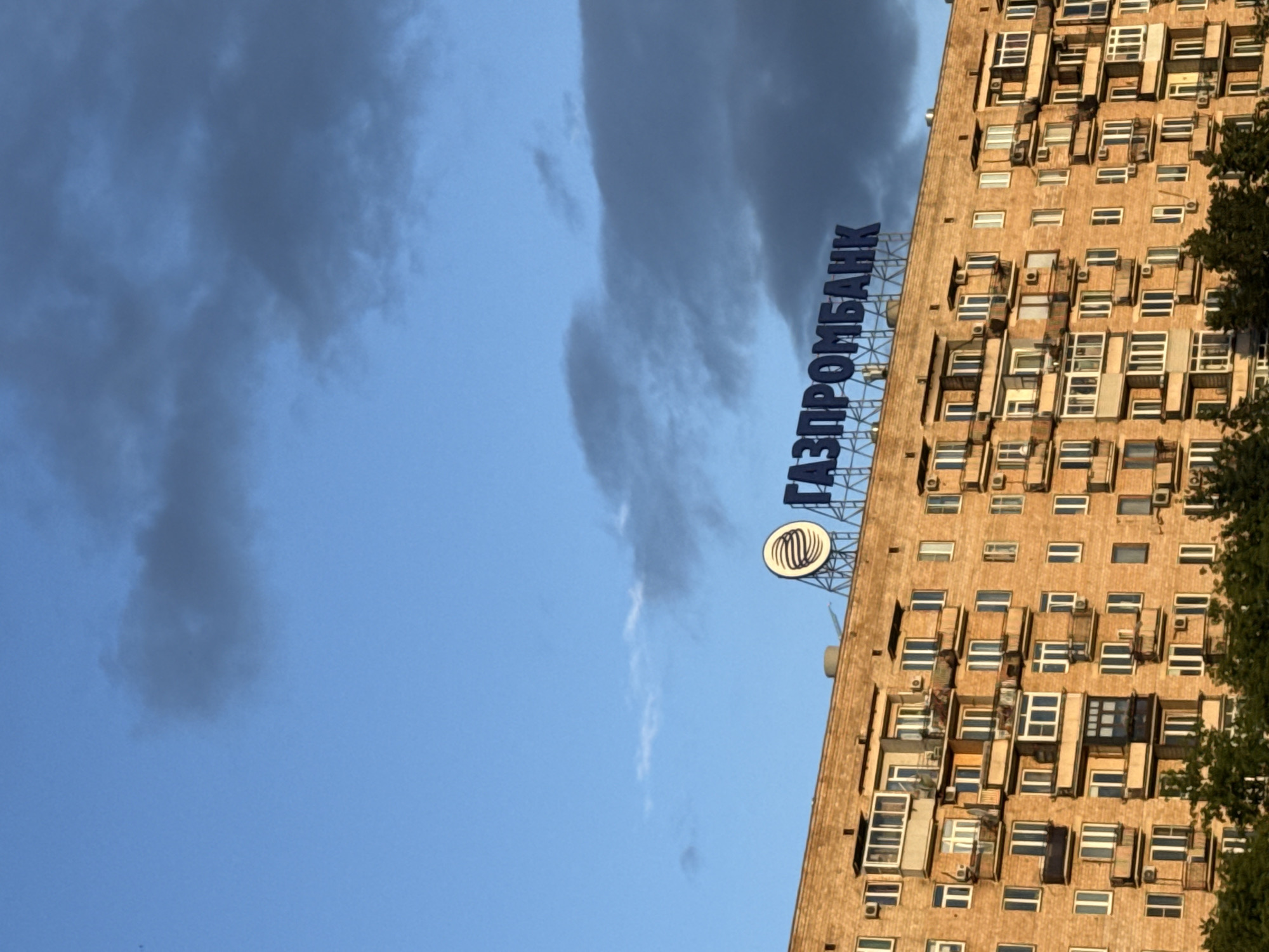 The image shows a brick apartment building in Moscow, Russia, with a large sign on top that reads "ГАЗПРОМБАНК". The sky is blue with some dark clouds. The building has many windows and balconies. There are trees in the foreground.