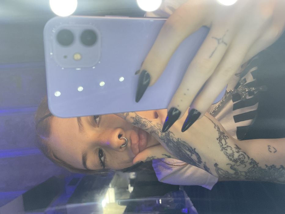 A young female individual is depicted in a mirror selfie, captured indoors. The subject has reddish-brown hair, light skin, and is looking towards the camera with a "duck face" expression, featuring pursed lips and prominent eyelashes. A silver septum ring is visible. Her left hand, adorned with intricate black ink scrollwork tattoos extending from the forearm to the hand, and a small heart tattoo on the lower forearm, supports her cheek. Her right hand, exhibiting long black pointed nails and small dot and crucifix-like tattoos on the fingers, holds a light purple mobile phone. The phone's dual rear cameras are visible, reflecting in the mirror. She is wearing a light-colored short-sleeved top, and a silver chain with a detailed cross pendant is partially visible around her neck. The background is dark and indistinct, with a discernible dark blue/purple ambient light. On the far left, a blurred glass bottle or container is present. The right side of the image features reflections of multiple bright, circular lights, indicating the use of a lighted vanity mirror. The precise location within the city of Buenos Aires, Argentina, cannot be determined from the visual evidence provided.