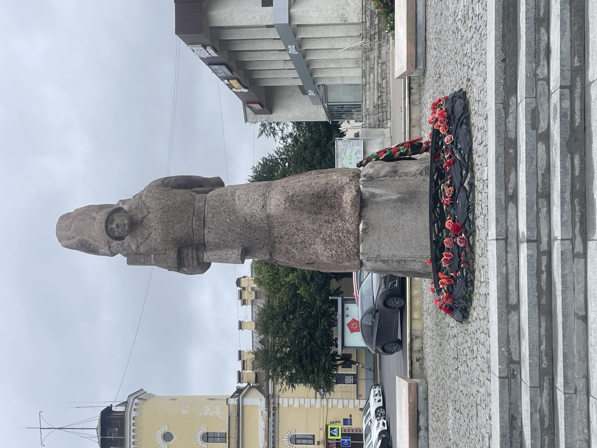 A monument of a soldier stands in Gatchina, Russia, with a wreath of red flowers at its base. The soldier is depicted wearing a helmet and holding an object in its left hand. The monument is made of rough-hewn stone and stands on a granite pedestal. In the background, a yellow building with a tower is visible, along with a modern building with columns. Several cars are parked in front of the buildings. The ground is paved with cobblestones, and there are steps leading up to the monument. The sky is overcast, suggesting a cloudy day.