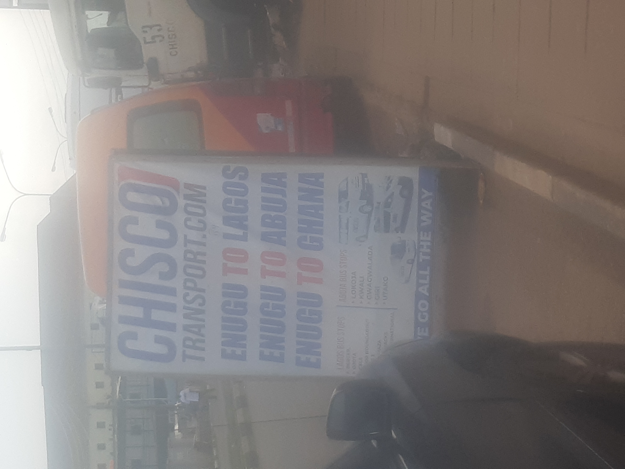 The image shows a sign for "CHISCO TRANSPORT.COM" which states routes such as "ENUGU TO LAGOS", "ENUGU TO ABUJA", and "ENUGU TO GHANA". The sign also lists bus stops for Lagos and Abuja. In the background, there are several vehicles, including what appear to be buses and trucks, suggesting this is a transportation hub or terminal. The environment appears to be an urban setting, possibly a busy street or a depot, with overcast weather conditions indicated by the hazy sky. The sign is positioned outdoors, on a sidewalk or roadside, and a portion of a black vehicle is visible in the foreground on the left. The sign is slightly tilted, and some parts are obscured by glare. The overall impression is that of a functional advertisement for a transport company operating within and beyond Enugu, Nigeria.