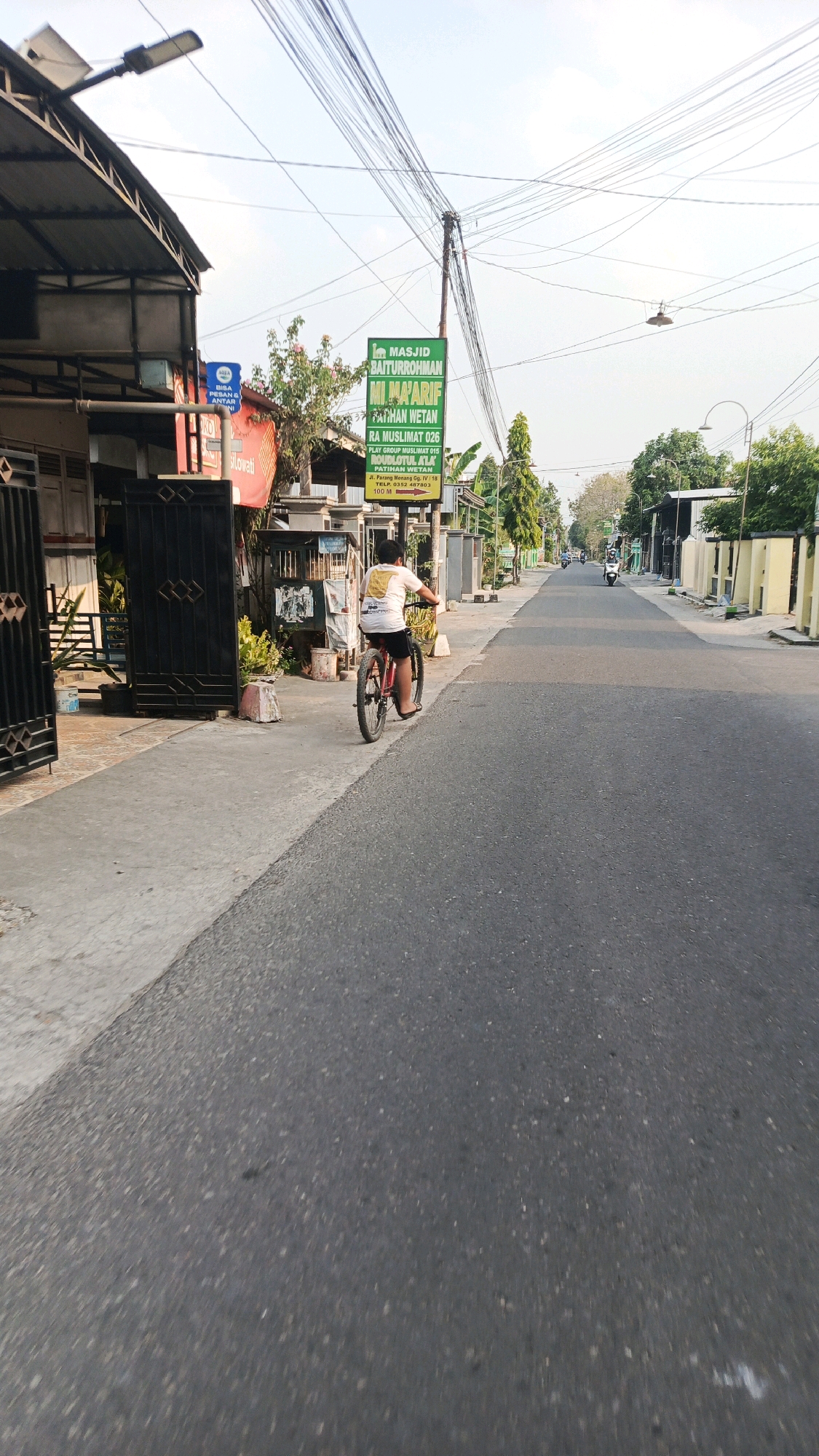 A young person rides a bicycle down a paved road in Patihan Wetan, Indonesia. The person is wearing a white t-shirt with a design on the back and black shorts, and is facing away from the camera. The road is lined with buildings on the left and right, and there are trees and power lines visible. A green sign for MASJID BAITURROHMAN is posted on the left side of the road. The sky is clear and the sun is shining.