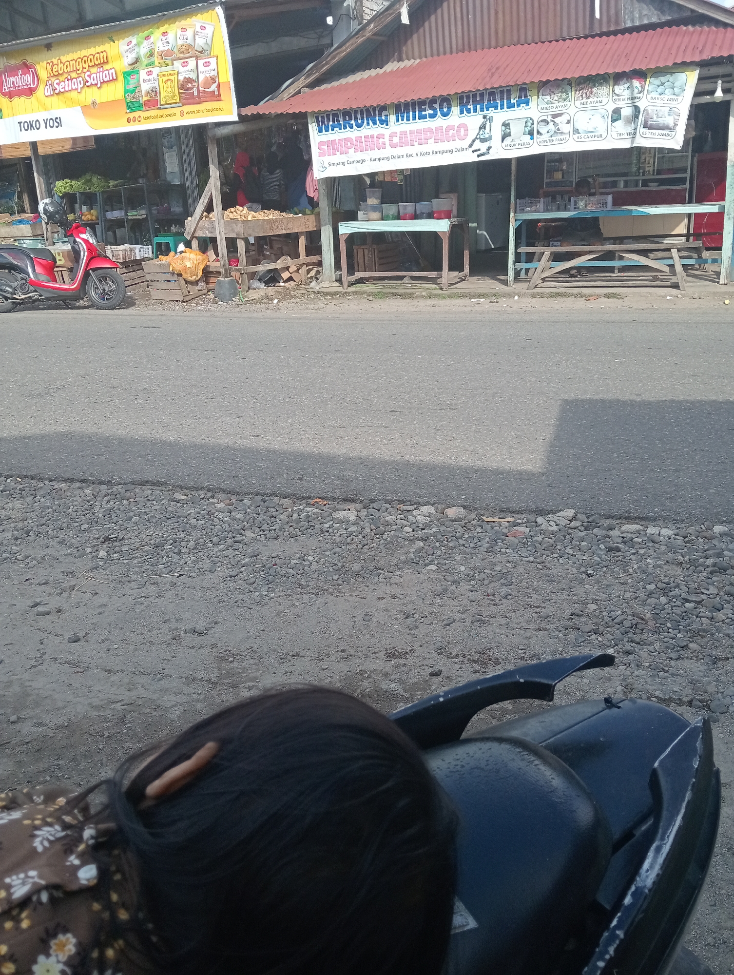 The image depicts a street scene in Campago, Indonesia. In the foreground, the back of a person's head with dark hair and a brown floral shirt is visible, resting on the seat of a black motorcycle. The motorcycle is parked on a gravelly surface next to a paved road.
Further back, a row of roadside stalls lines the street. On the left, a red motorcycle is parked in front of a stall with a yellow banner that reads "Abrofood Kebanggaan di Setiap Sajian" and "TOKO YOSI". The banner also features product images and website addresses. The stall appears to sell various goods, with shelves visible behind the counter.
To the right, another stall has a red corrugated metal roof and a banner that says "WARUNG MIESO KHAILA" and "SIMPANG CAMPAGO". This stall also displays images of food and drink items. Inside the stall, a person is seated behind a counter, and various items are displayed on shelves and tables, including what appear to be containers of food. Wooden benches are set up in front of this stall for seating. The environment suggests a bustling local marketplace.