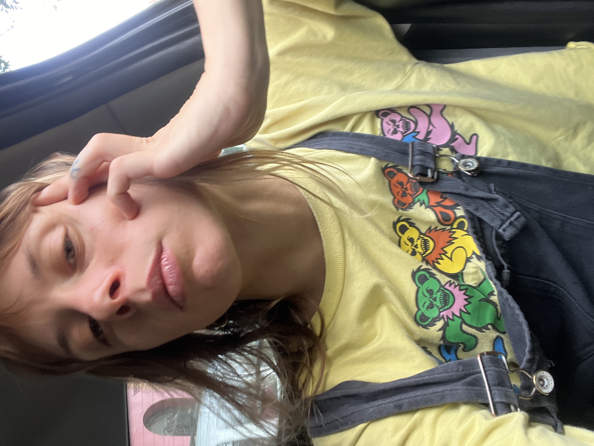 A person is in a vehicle, possibly a car, wearing a yellow t-shirt with a Grateful Dead dancing bears motif and gray denim overalls. The person is touching their face with their right hand. The background shows glimpses of an urban environment, potentially a street or building exterior, although specific details are unclear. There is no clear indication of the location being Buenos Aires, Argentina, or any specific city.