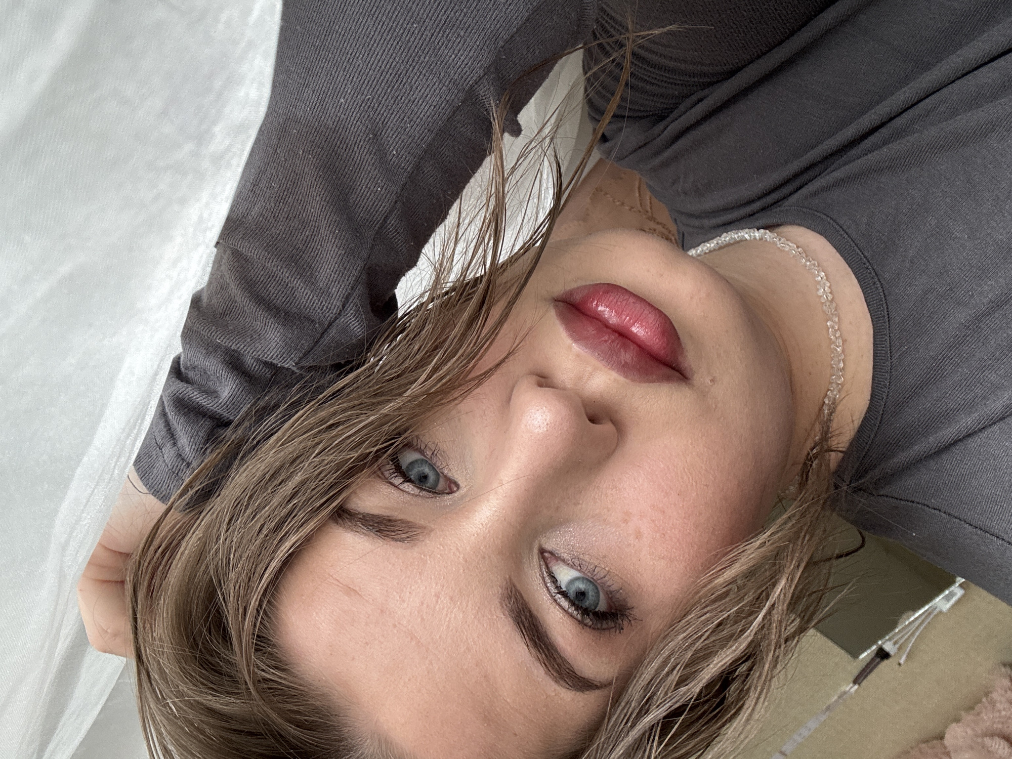 A close-up, angled shot of a woman with fair skin and light brown hair. She is wearing a gray long-sleeved shirt and a delicate silver necklace. Her eyes are light blue, and her lips have a glossy, dark pink hue. Her hair appears damp or styled with product. The background includes a white fabric, possibly a sheet, and a glimpse of a neutral-toned floor with indistinct objects. The location in the city of Chaikovsky, Russia is not evident from the image.