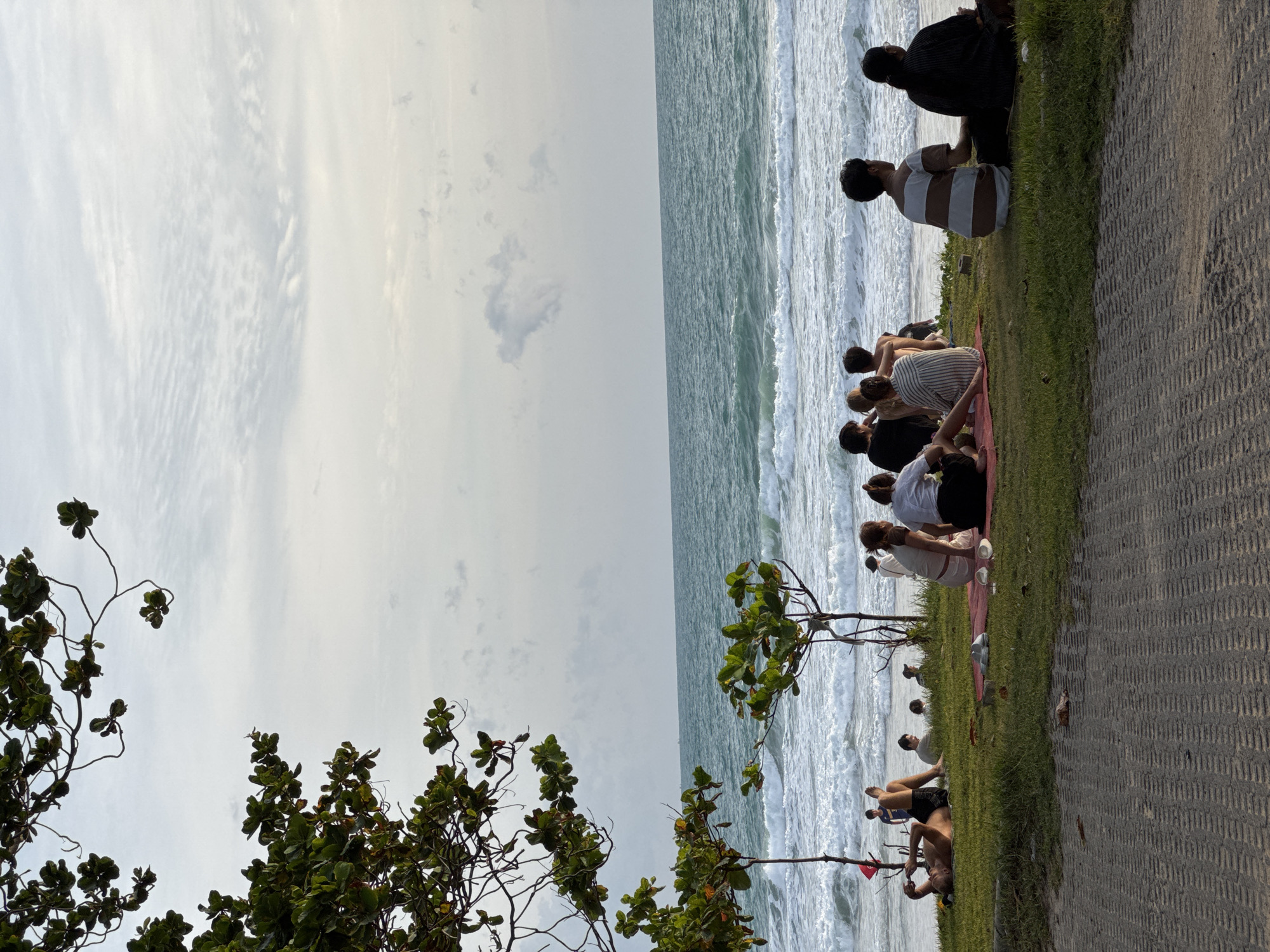 A group of people are sitting on the grass near the beach in Choeng Thale, Thailand, facing the ocean. The sky is overcast with light clouds. The ocean has moderate waves and a sandy shore. There are trees with green leaves on the left and in the middle of the frame. In the foreground, there is a paved walkway. One person is lying on the grass on their back, looking at their phone. Several individuals are seated on mats, while others are directly on the grass.
