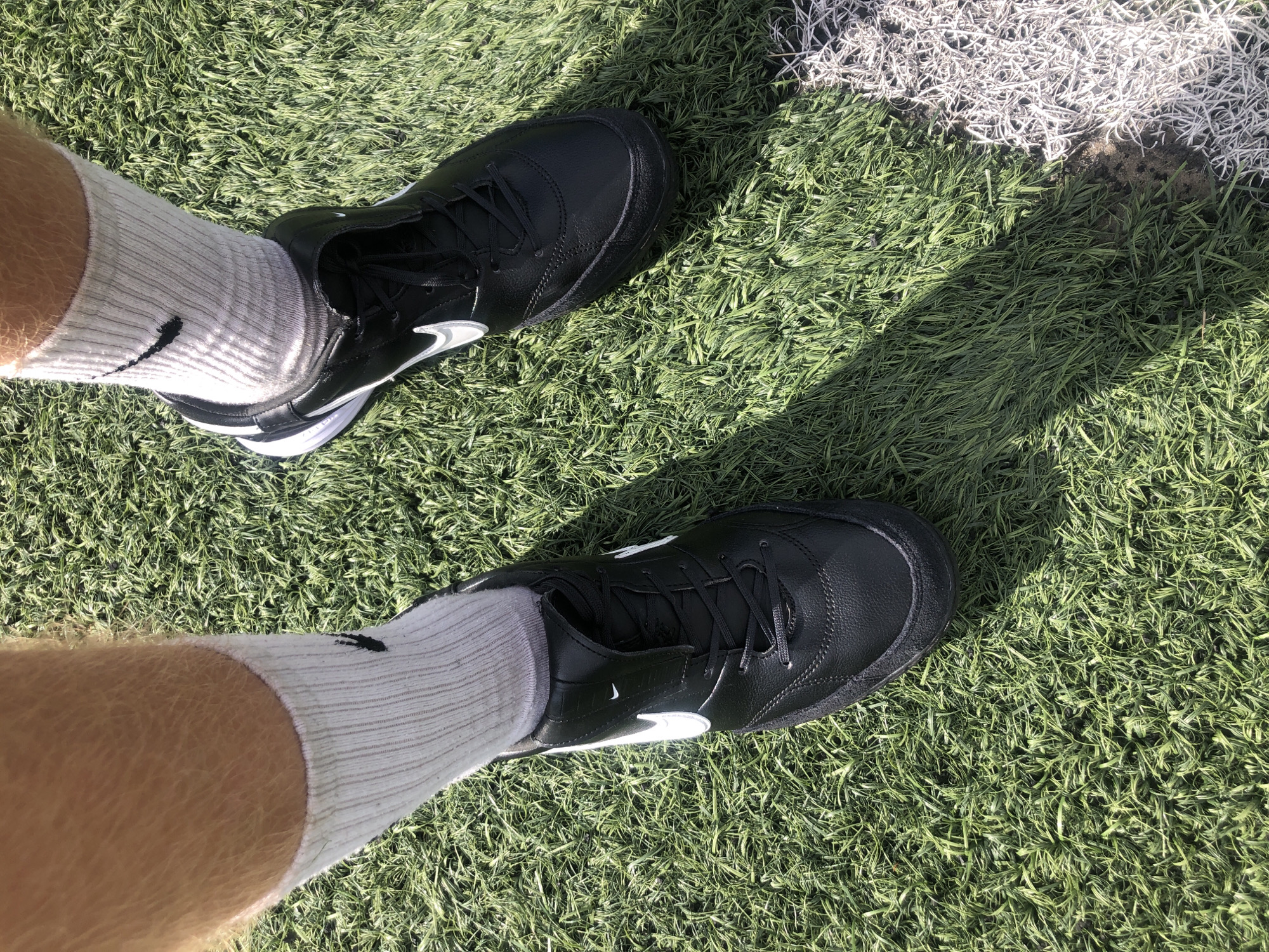The image shows a pair of black Nike soccer cleats with white Nike swooshes on a synthetic turf field. The cleats are worn by a person with light-colored, hairy legs wearing white athletic socks. The turf is green with a shadow cast across it from the sun. A white painted line is visible on the right side of the image, marking the boundary of the field. The scene is likely an outdoor soccer field in Dnipro, Ukraine.