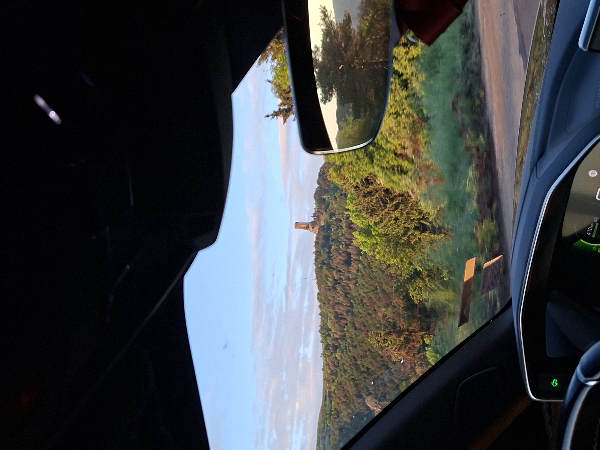 The image provides a driver's or front passenger's perspective from inside a modern Peugeot vehicle, looking out through the front windshield at a scenic landscape in Charkovo, Bulgaria. The time of day appears to be either late afternoon or early morning, characterized by the warm, soft light illuminating a dense, forested hillside under a mostly clear, bright sky with scattered clouds.
A prominent feature in the distant landscape is a tall, stone-colored tower-like structure or ruin, standing distinctly above the treeline on the hilltop. A light-colored road is visible running alongside the forest, suggesting travel through this natural setting.
Inside the car, the dark dashboard and vehicle controls are partially visible on the right side of the frame. A digital display shows "410 km," likely indicating remaining driving range, along with a green downward-pointing arrow symbol. The word "PEUGEOT" is faintly discernible on a console. The rearview mirror, positioned centrally at the top, reflects more of the surrounding trees and sky. Reflections on the windshield include a red object, possibly part of the car's interior or a sun visor, and a sticker with faint, illegible text on the lower right. The overall scene conveys a sense of calm travel through a picturesque natural environment.