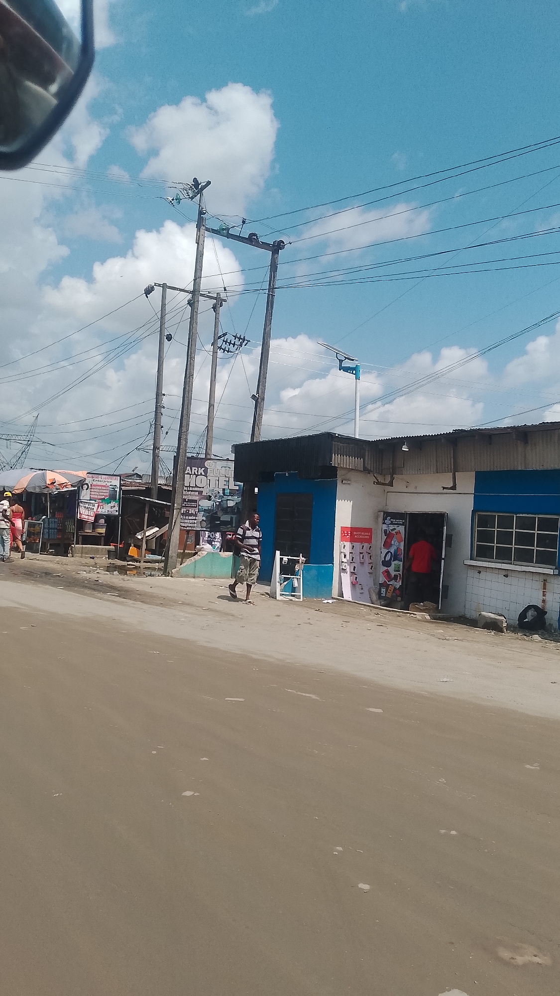 The image depicts a street scene in Effurun, Nigeria, with a sandy road leading to a row of low buildings and commercial establishments. Overhead power lines crisscross the sky, which is partly cloudy. A man is walking along the side of the road, near a blue and white storefront advertising phone accessories. To the left, there are open-air stalls with goods displayed, and people are present in that area. The buildings have a somewhat weathered appearance, and the scene has a general atmosphere of a typical urban street in a developing country.