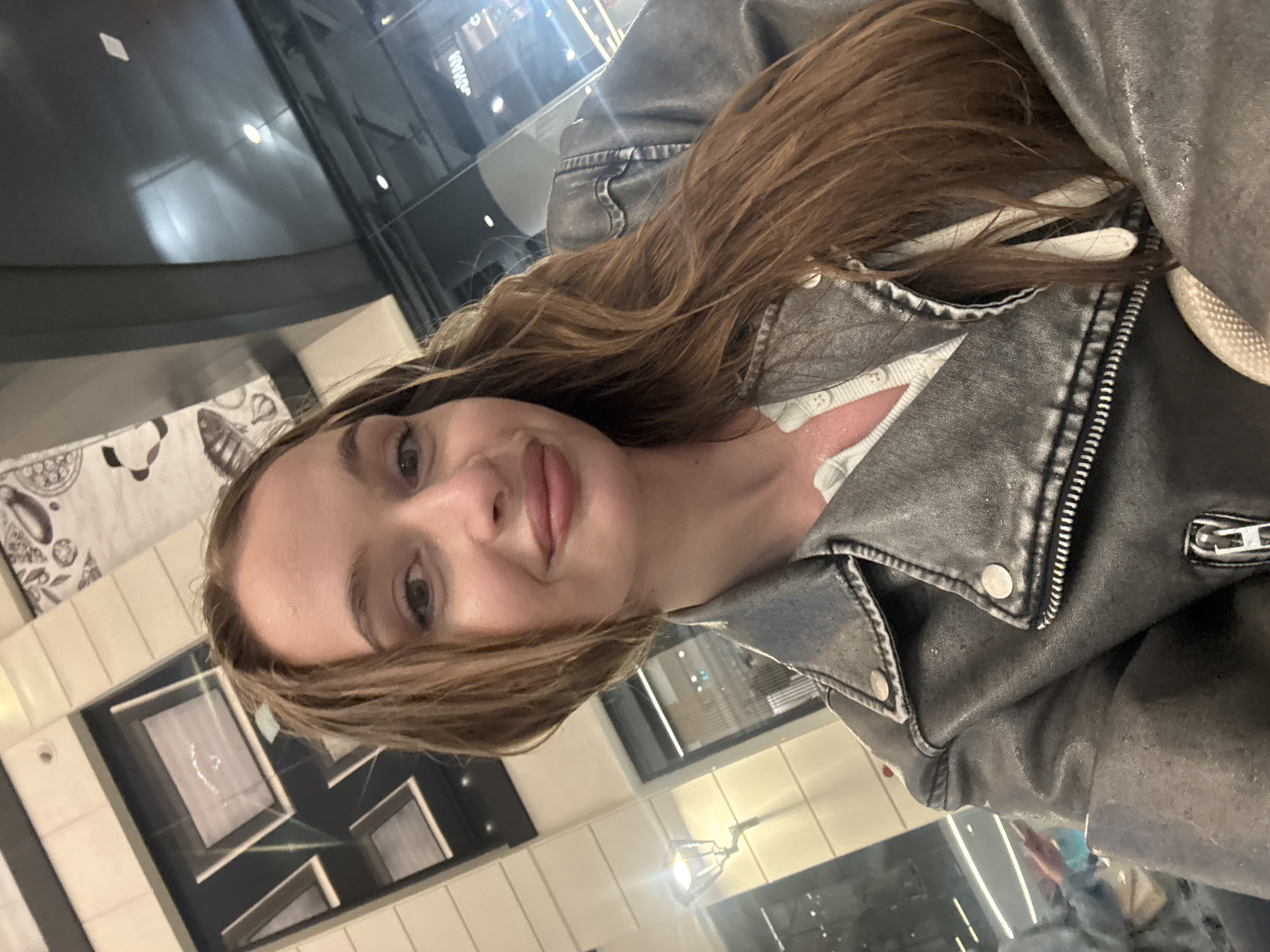 A woman with long, brown hair and fair skin is taking a selfie. She is wearing a white top and a dark grey leather jacket. The background appears to be an indoor location, possibly a restaurant or cafe in a modern-looking building. Reflected in a glass surface are lights and building features, suggesting an urban environment. The exact location within Belgrade, Serbia, cannot be determined definitively from the image alone.