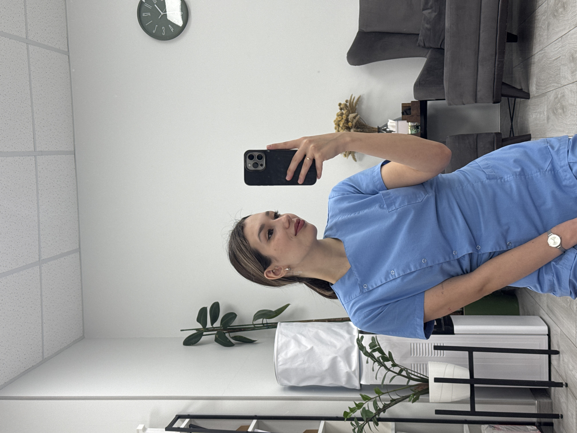 The image displays a young woman taking a selfie in an indoor environment, likely a professional setting such as a clinic, office, or waiting room. She is positioned slightly to the right of the frame, holding a black smartphone with a triple camera array in her right hand, directed towards the camera as if capturing her reflection. She has dark brown hair, styled with a side part, and looks directly towards the phone with a neutral and composed expression. She is dressed in a light blue, short-sleeved scrub-style top with buttons down the front. A silver watch adorns her left wrist, and a simple gold band ring is visible on her right ring finger.
The background features clean white walls and a white acoustic ceiling with a visible grid pattern. To the right, part of a wall with light-colored wooden paneling or a wooden floor is visible. In the upper right corner, two dark grey, modern armchairs or chairs are present, partially obscuring a small dark wooden table or cabinet which holds a dried floral arrangement. Closer to the foreground on the left, there's a black-framed shelving unit with white shelves, displaying various items including two potted plants – one with large dark green leaves and another with smaller green leaves. A round, dark grey wall clock with minimalist markings, including stylized numbers like '3' and '9', is mounted on the upper left wall, indicating the time is approximately 12:05. The lighting in the room is bright and even. There is no visible text apart from the stylized numbers on the clock face.