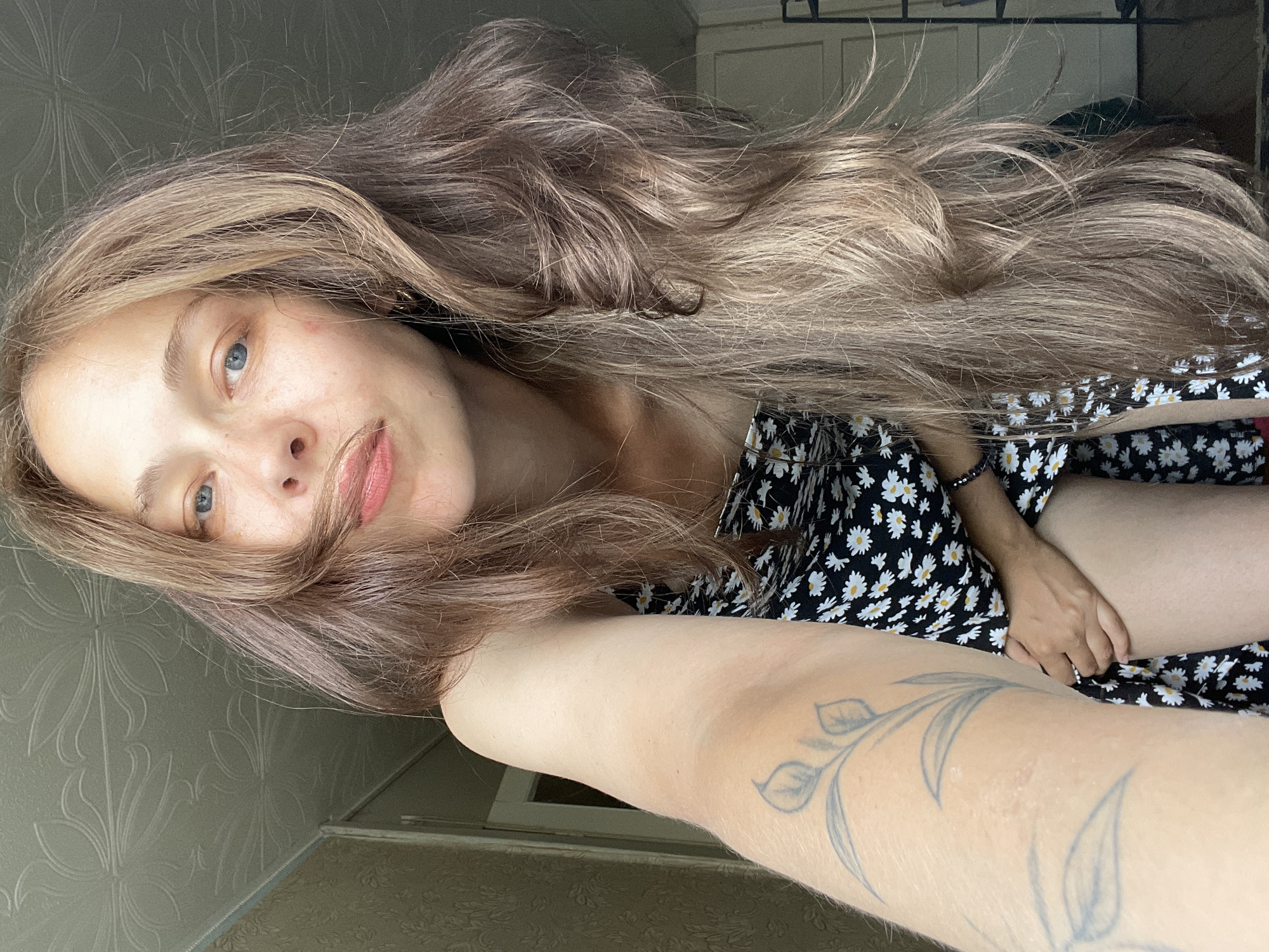 A young woman with long, wavy, brown and blonde hair is shown in a selfie from the chest up. She is wearing a black dress with a white daisy pattern. She has blue eyes and is looking at the camera. Her left arm is raised, holding the camera, and a tattoo of a plant with leaves and a flower is visible on her forearm. The background appears to be a room with textured wallpaper and a patterned ceiling. The lighting suggests it is daytime.
