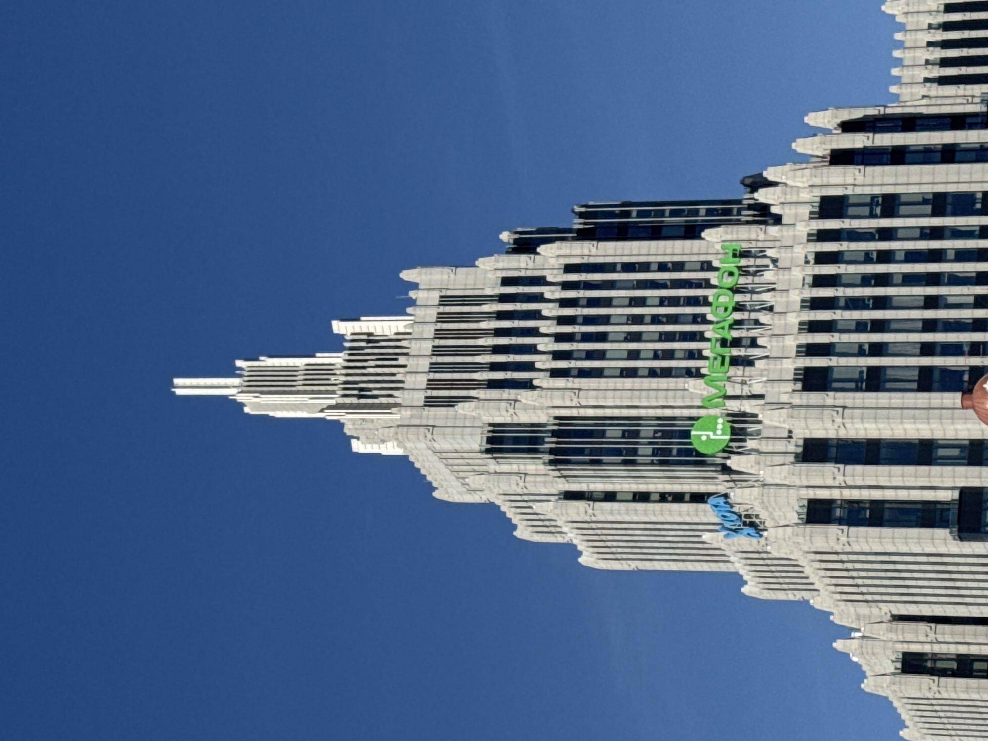 The image displays a towering, multi-story building against a clear, vibrant blue sky. The building, likely a skyscraper, exhibits a distinctive stepped or tiered architectural style, with numerous setbacks as it ascends. Its facade is light-colored, possibly white or light grey stone/concrete, contrasting with dark, rectangular windows arranged in horizontal bands. The overall impression is one of a grand and imposing structure in an urban setting.
No human activity is visible, making the building the sole subject. The bright, cloudless sky indicates clear weather conditions and daytime, likely mid-day due to the uniform illumination and lack of long shadows.
Prominently displayed on one of the building's upper sections are large, bright green letters spelling "WEWORK" vertically. Below this text, a green circular logo, also associated with the WeWork brand, is visible. Further down the facade, partially obscured, are blue letters that spell out "VTB". This suggests the building houses offices for both WeWork and VTB. Given the provided context, this building is located in Moscow, Russia.