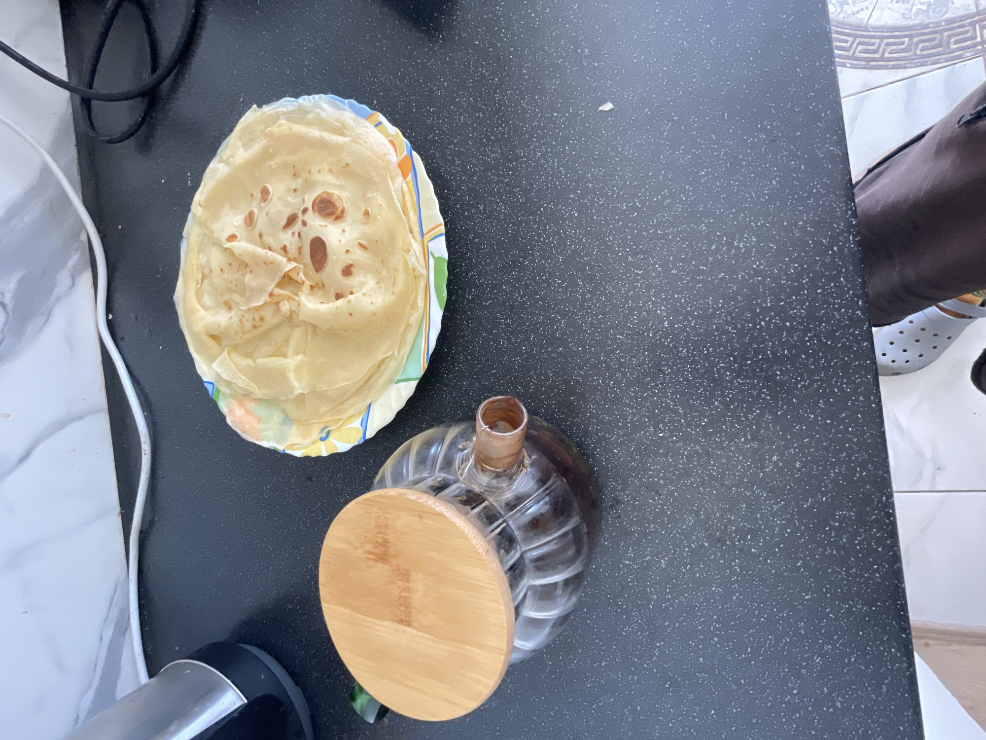 A high-angle shot captures a breakfast scene on a speckled black countertop in Bol'shoe Zaborod'e, Russia. In the foreground, a clear glass carafe filled with a dark liquid, possibly coffee or tea, is topped with a round, light-brown wooden lid. To the right of the carafe, a stack of thin, golden-brown crepes rests on a plate with a colorful rim featuring floral patterns. The background shows a white, marble-patterned backsplash and coiled black and white electrical cords. The scene is illuminated by overhead lighting, casting subtle shadows.