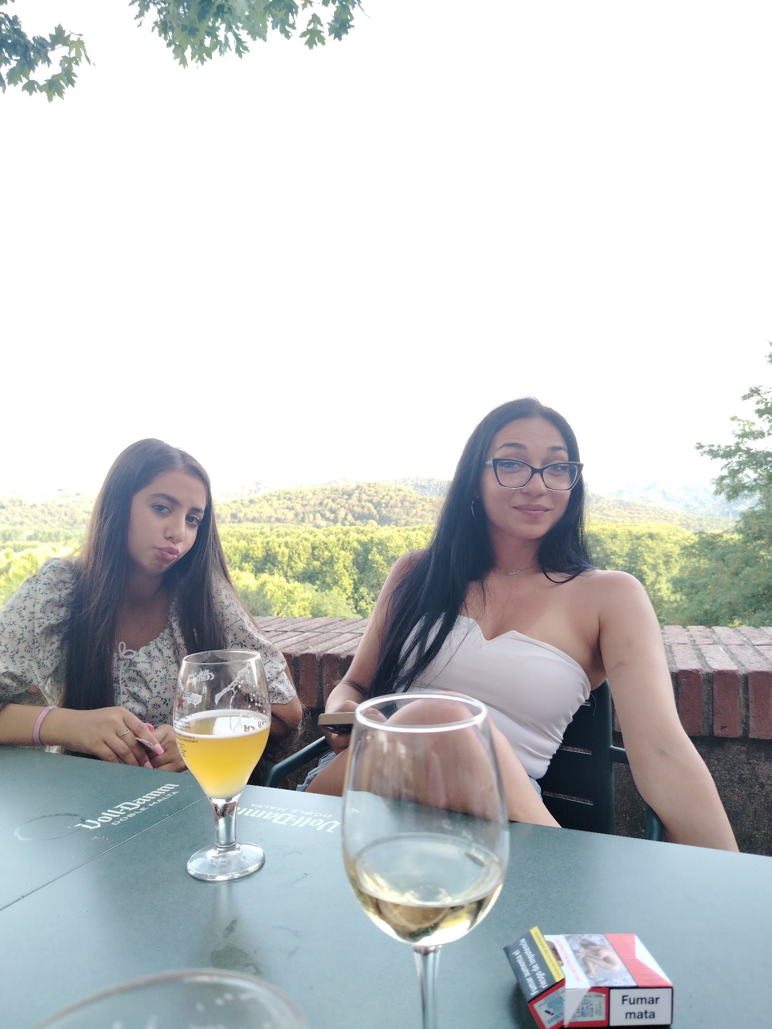 Two young women are seated at a table outdoors, with a scenic view of a forested hillside in the background. The woman on the left has long dark hair and is wearing a floral print top. She is looking at the camera with her lips pursed. In front of her is a glass of beer. The woman on the right has long dark hair and is wearing a white strapless top and glasses. She is smiling and looking at the camera. In front of her is a glass of white wine and a pack of cigarettes. The table is green and has a circular logo with text that appears to be in Spanish. A low brick wall runs along the edge of the seating area. The foliage of trees is visible in the upper part of the image. This location appears to be a terrace or restaurant overlooking a valley, consistent with a view from the castle walls in Hostalric, Spain.