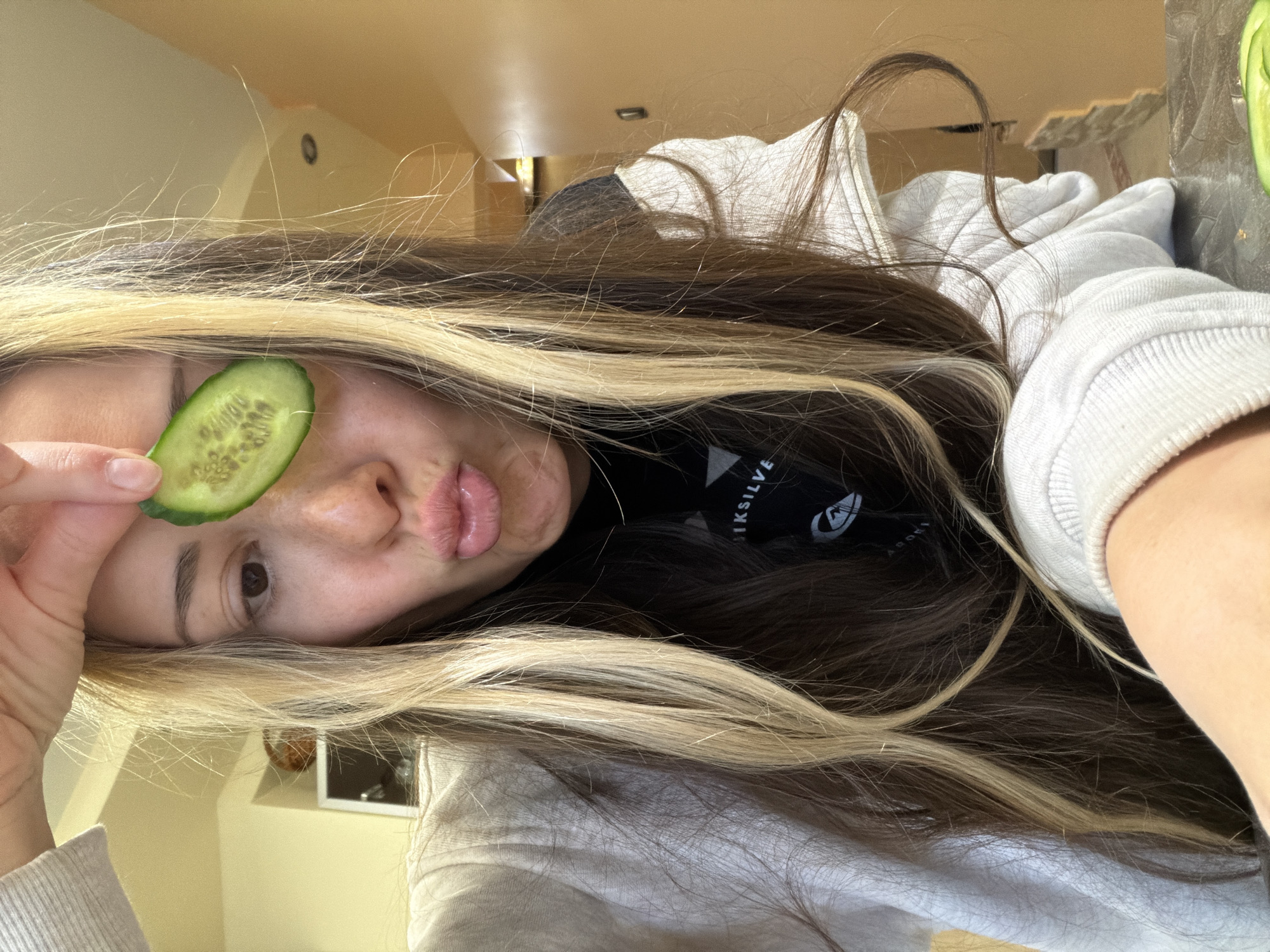 A young woman is holding a slice of cucumber over her left eye and pouting her lips. She has long brown hair with blonde highlights. She's wearing a white hoodie over a black top with a white logo. The background is blurry but appears to be a domestic interior. The location is not discernible.