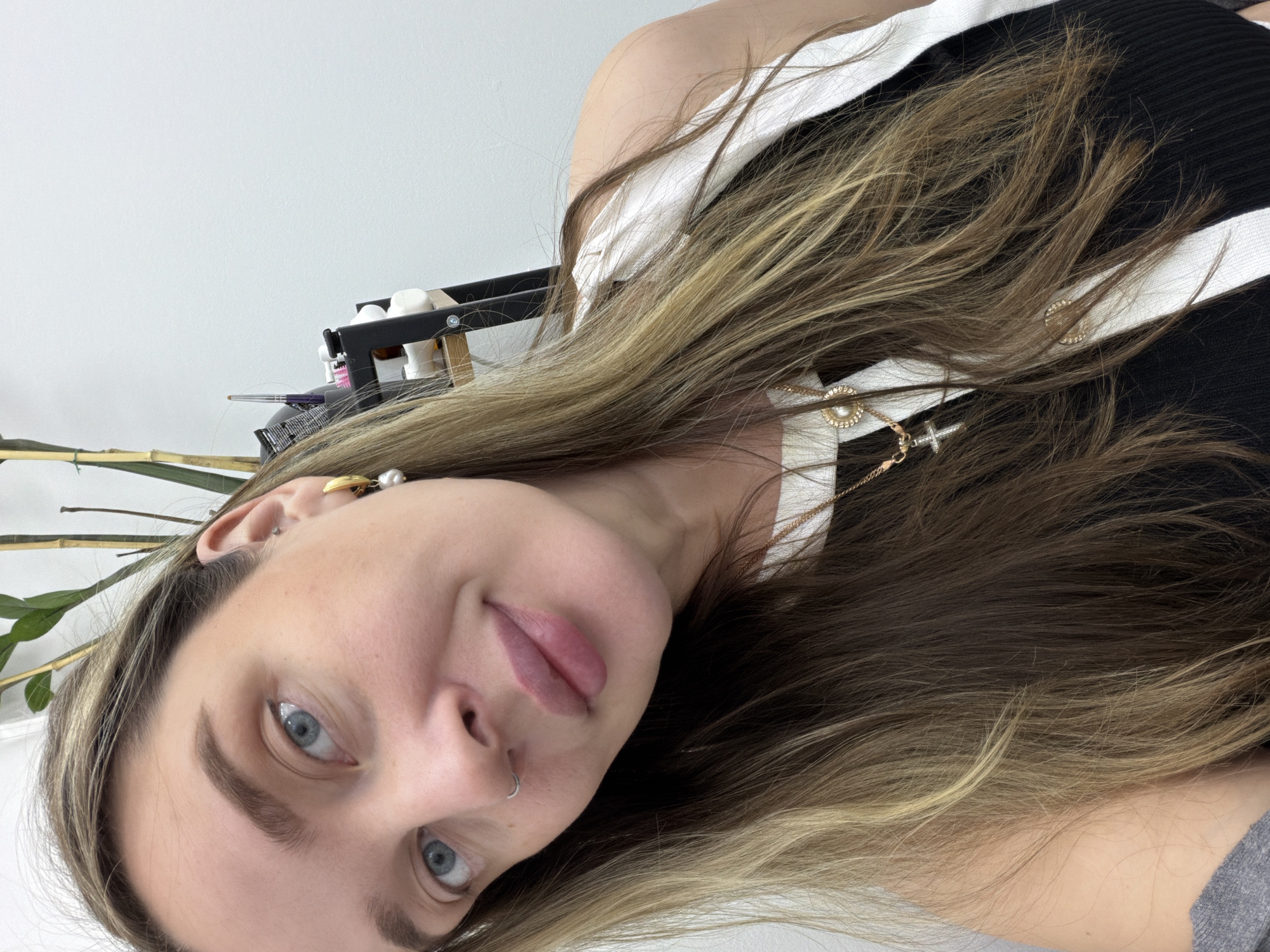 A young woman with light skin and blue eyes is looking directly at the camera. She has long, brown and blonde highlighted hair. She is wearing a black and white striped top and gold jewelry, including earrings, a necklace, and a nose ring. The background includes a white wall, some greenery, and a shelf with miscellaneous items. There is no indication of the location being Moscow, Russia.