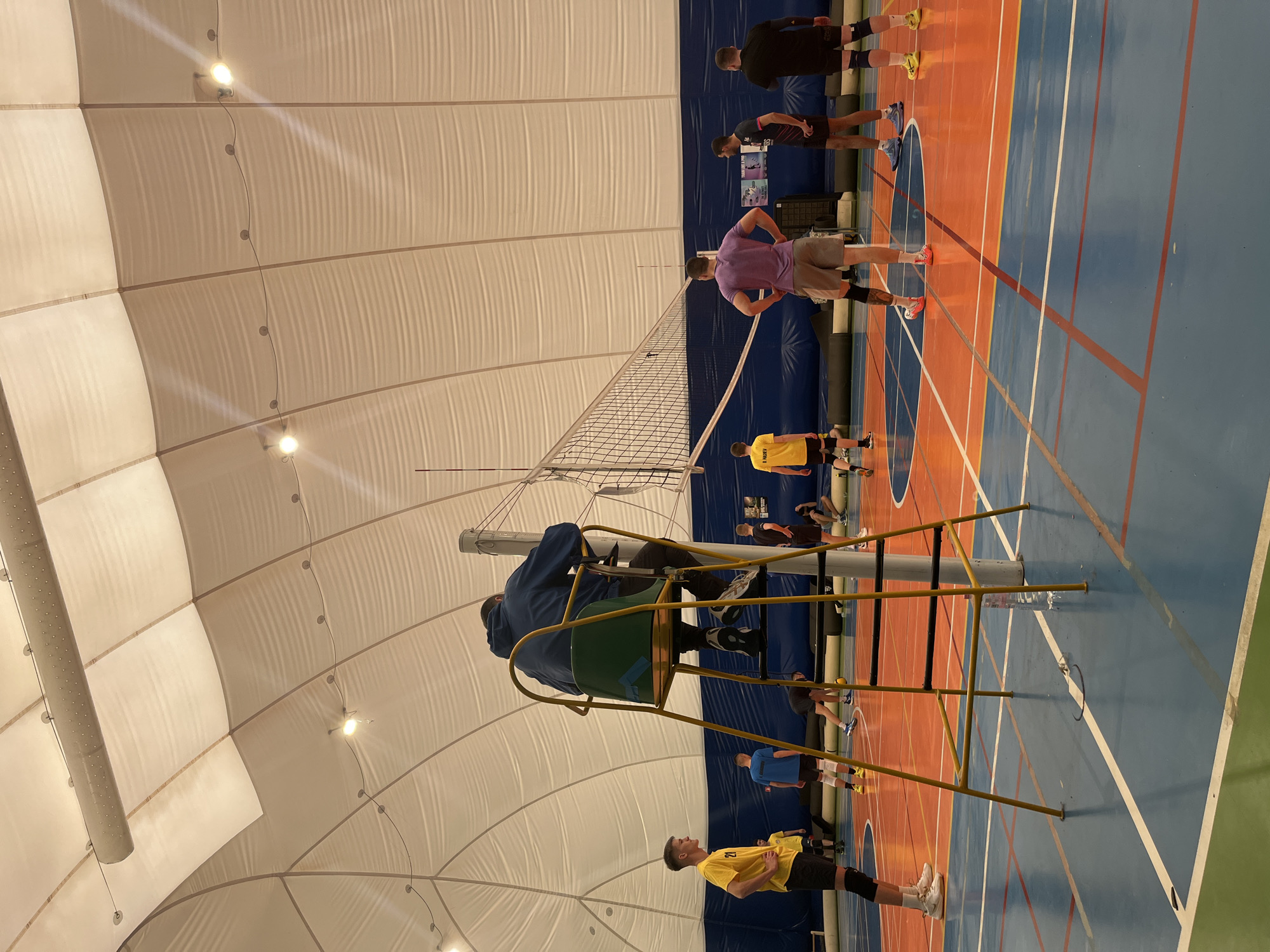 The image shows a volleyball game in an indoor sports facility with a high, domed ceiling. The court is marked with blue and orange lines. A volleyball net stands in the center, with a referee sitting on a high chair next to it. Several people, mostly young men, are standing on the court, some in athletic attire. The building has rounded segments composing a large, spherical roof. This photo seems to be taken inside a sports hall. The location in the city of Sofia, Bulgaria is not known.