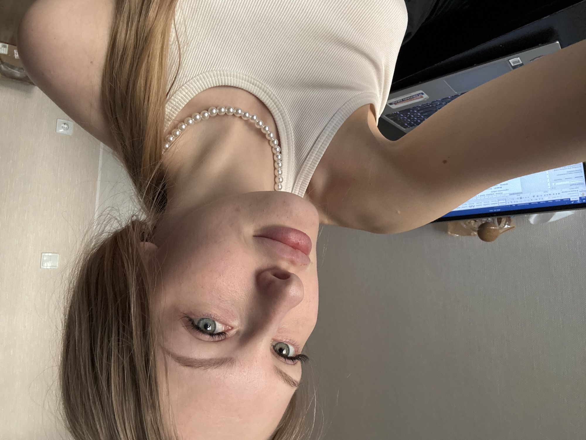 A portrait-oriented image, rotated 90 degrees counter-clockwise, captures a young woman from a slightly upward angle, likely a selfie. She has light brown hair parted on the side, striking blue-green eyes with prominent eyelashes, and light skin. Her lips are slightly parted, and she has a calm, perhaps neutral or contemplative expression. She is wearing a light-colored, ribbed tank top and a single strand pearl necklace.
The background to her right (top of the image as displayed) shows a light-colored wall with a white electrical outlet, consistent with European or Russian standards. To her left (bottom of the image as displayed) is a dark surface, likely a desk, with an open silver/gray laptop. A "Lenovo" brand sticker and an "Intel" logo are visible on the laptop. Beyond the laptop, a computer monitor displays what appears to be text from a document, though the content is unreadable. The setting suggests an indoor environment, possibly a home or office in Perm, Russia. A small, brown, possibly wooden, decorative object is barely visible near the bottom edge of the image, close to the monitor.