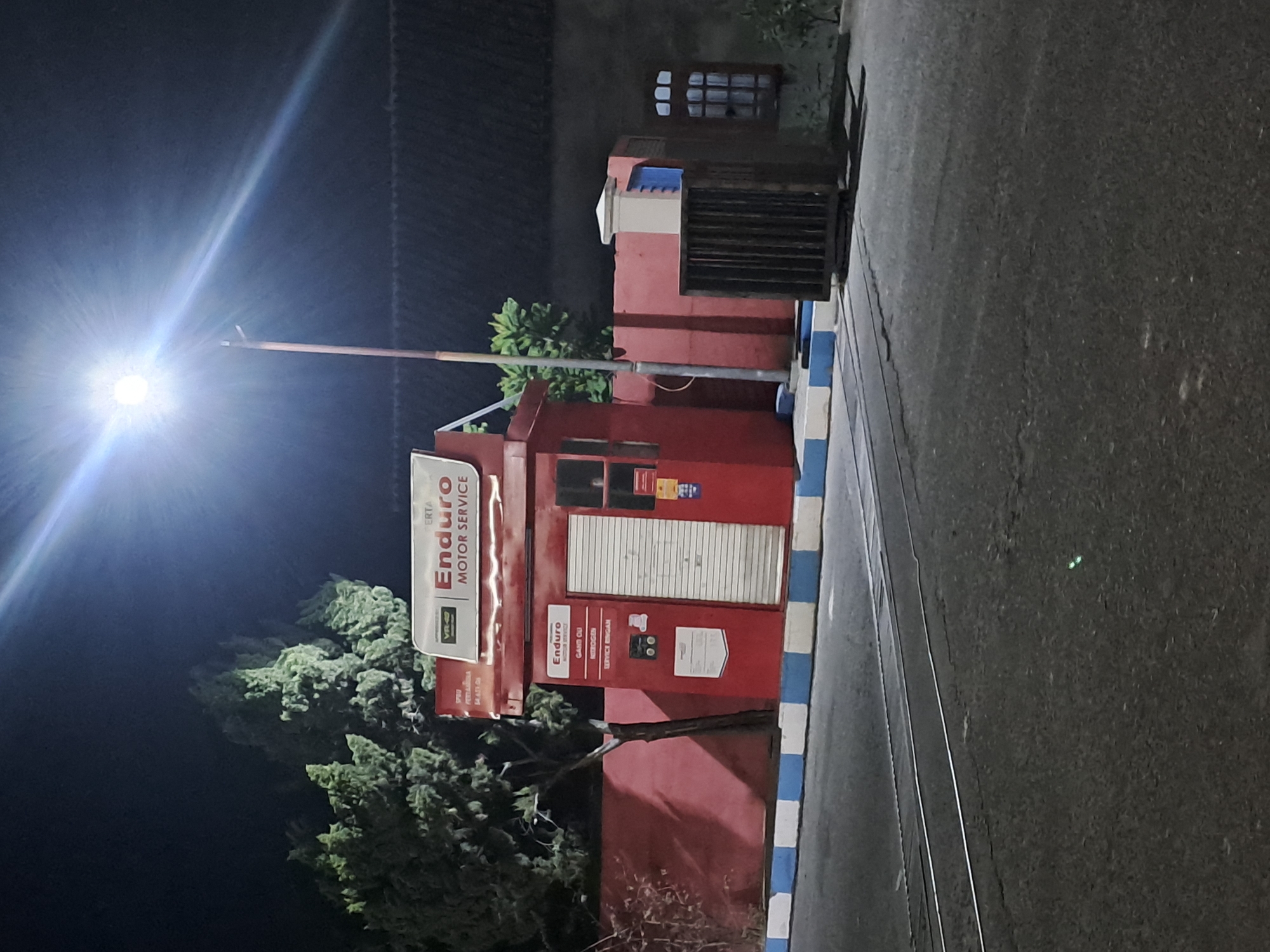 This image shows a red Enduro Motor Service station in Slambur, Indonesia at night. A bright street light illuminates the scene from above. The service station is a small, enclosed building with a red facade and a metal roller door. There is signage on the front of the building indicating "Enduro MOTOR SERVICE". Adjacent to the building, there is a red wall and a dark metal cage. In the foreground, there are train tracks running across the asphalt road. The area is dark, with shadows cast by the street light and the building. There are no people visible in the image.