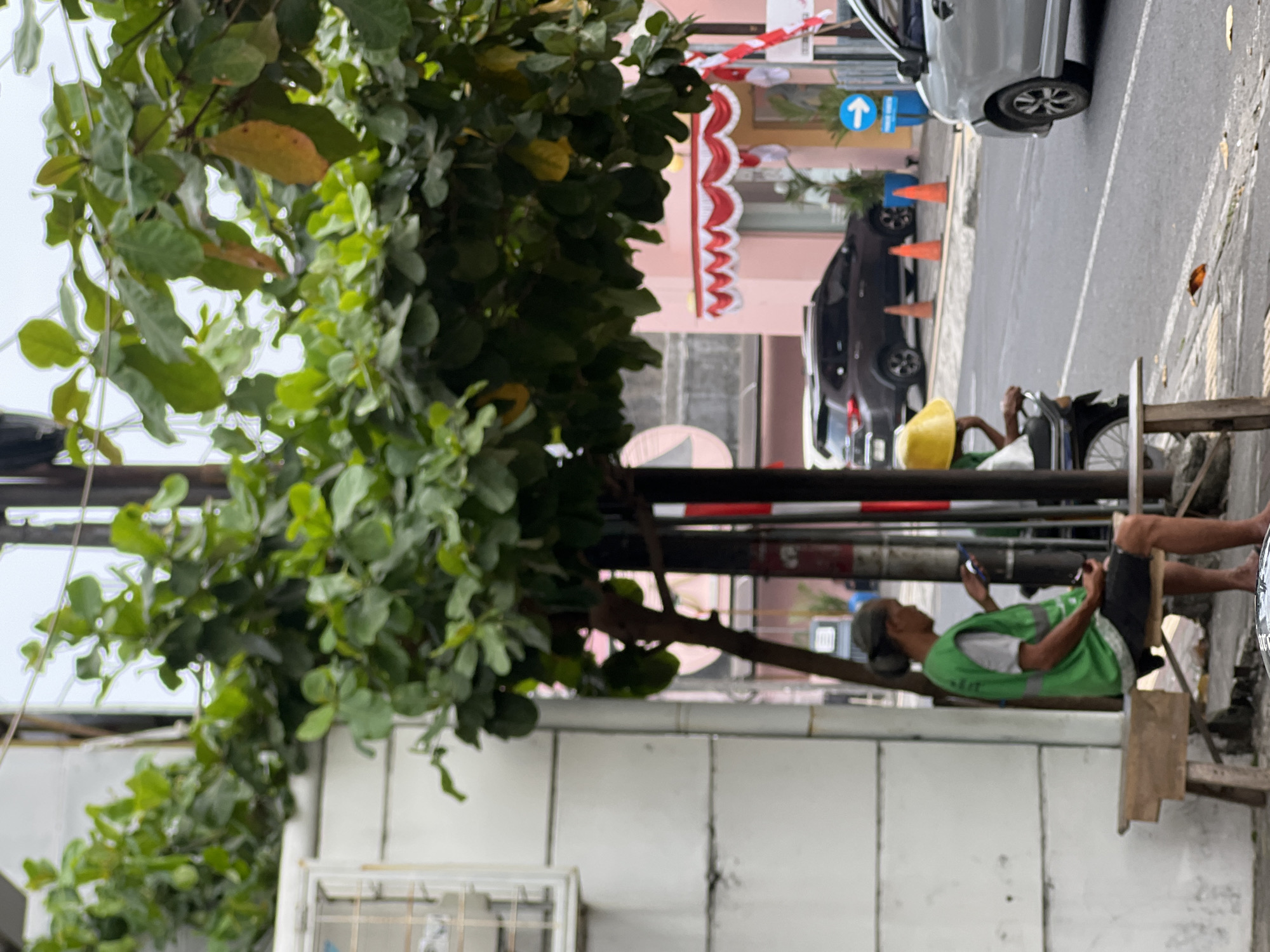 The image depicts a street scene in Yogyakarta, Indonesia. A man wearing a green vest and dark shorts sits on a wooden bench, looking at his mobile phone. To his right, another person wearing a yellow hat is partially visible, seated on a motorcycle. In the background, a dark-colored SUV is parked on the street, with traffic cones placed behind it. A light silver car is in motion on the road. The street is lined with buildings, one of which has a pink facade adorned with red and white decorative trim. Lush green foliage from a tree overhangs the scene, with some leaves appearing yellowed. An air conditioning unit is mounted on a white wall to the left of the frame. The environment suggests an outdoor urban setting, possibly in a commercial or residential area.